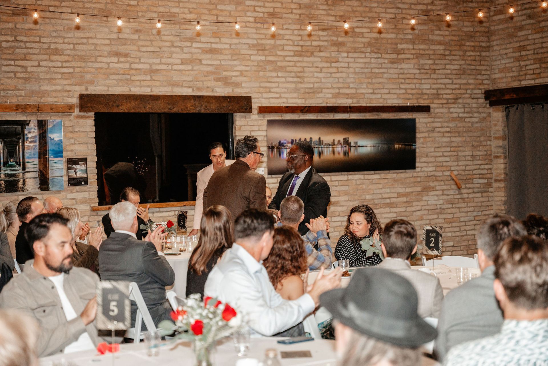 People at tables applaud as two men shake hands in a brick-walled room with string lights.