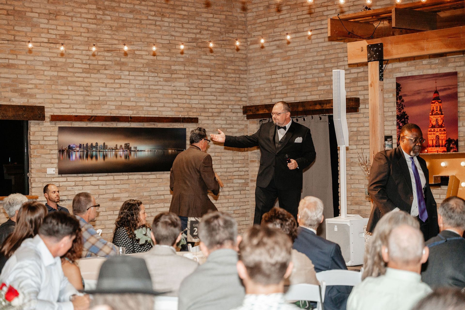 A man in a tuxedo gestures towards another man on a stage, speaking at an event with an audience.
