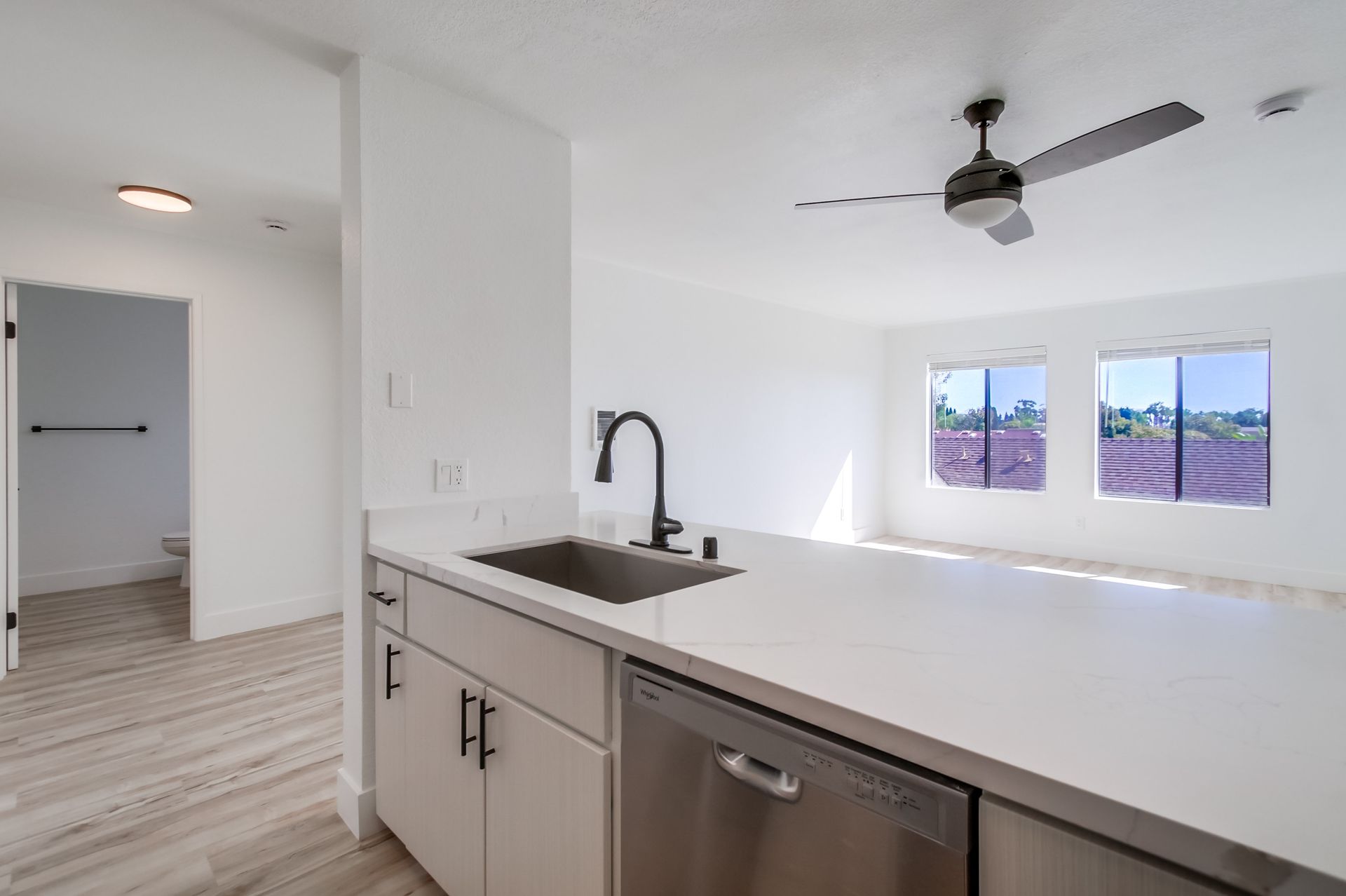 Bright white kitchen with a sink, counter, and view of a living room with a ceiling fan and windows.