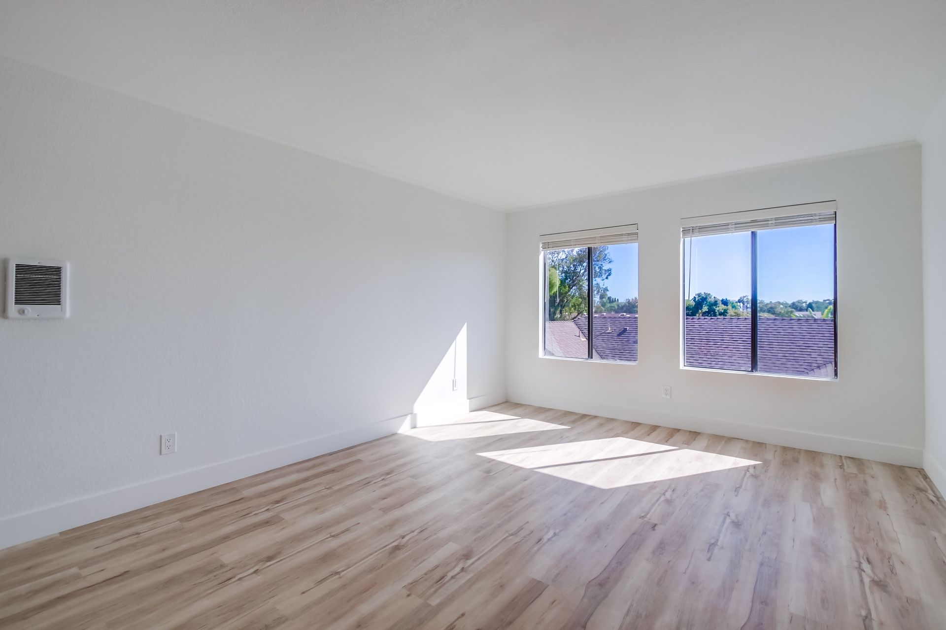 Empty room with wood-look flooring, white walls, and two windows. Sunlight streams in.