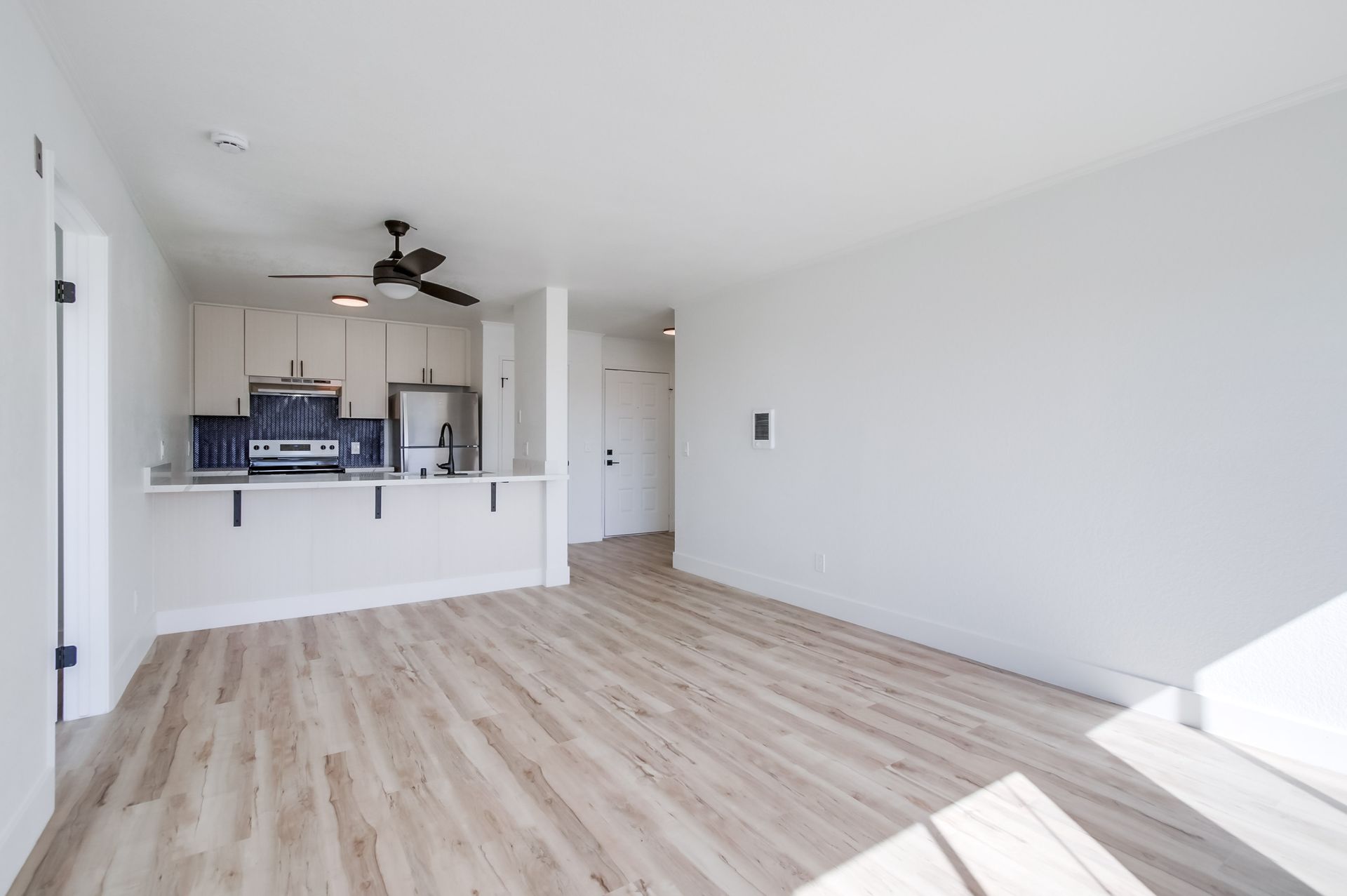 Empty, bright apartment with light wood floors, white walls, and a kitchen area.