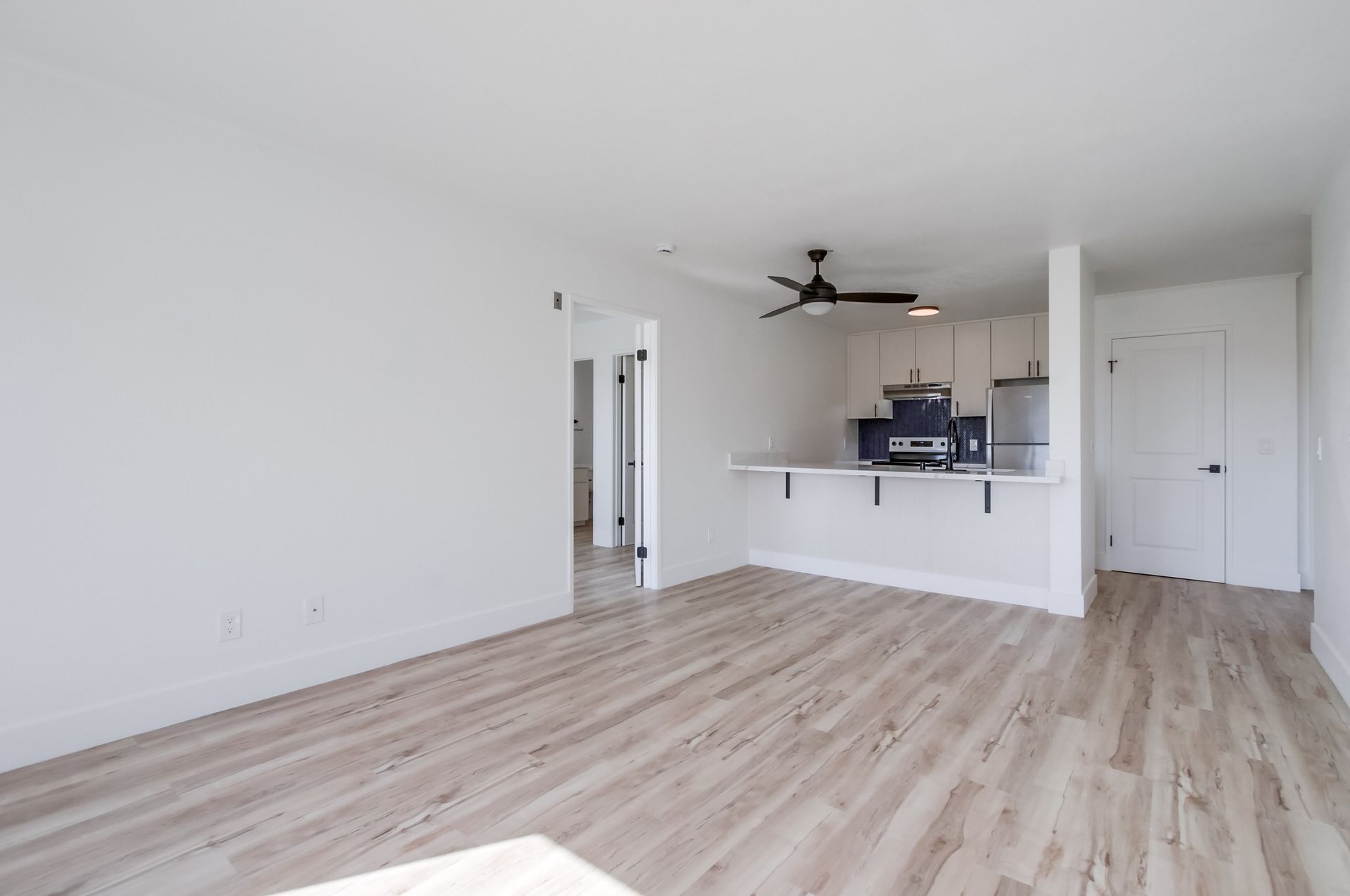 Empty living space with light wood floors and a small kitchen area.