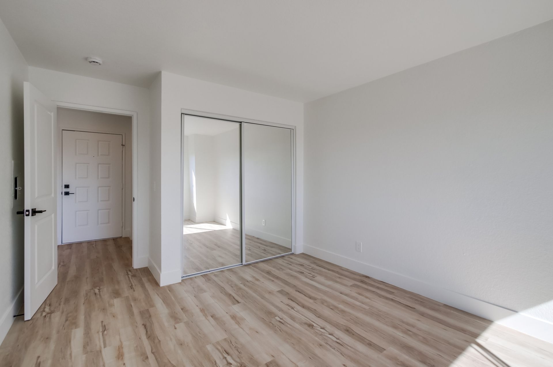 Empty bedroom with light wood floors, mirrored closet doors, and an open doorway.