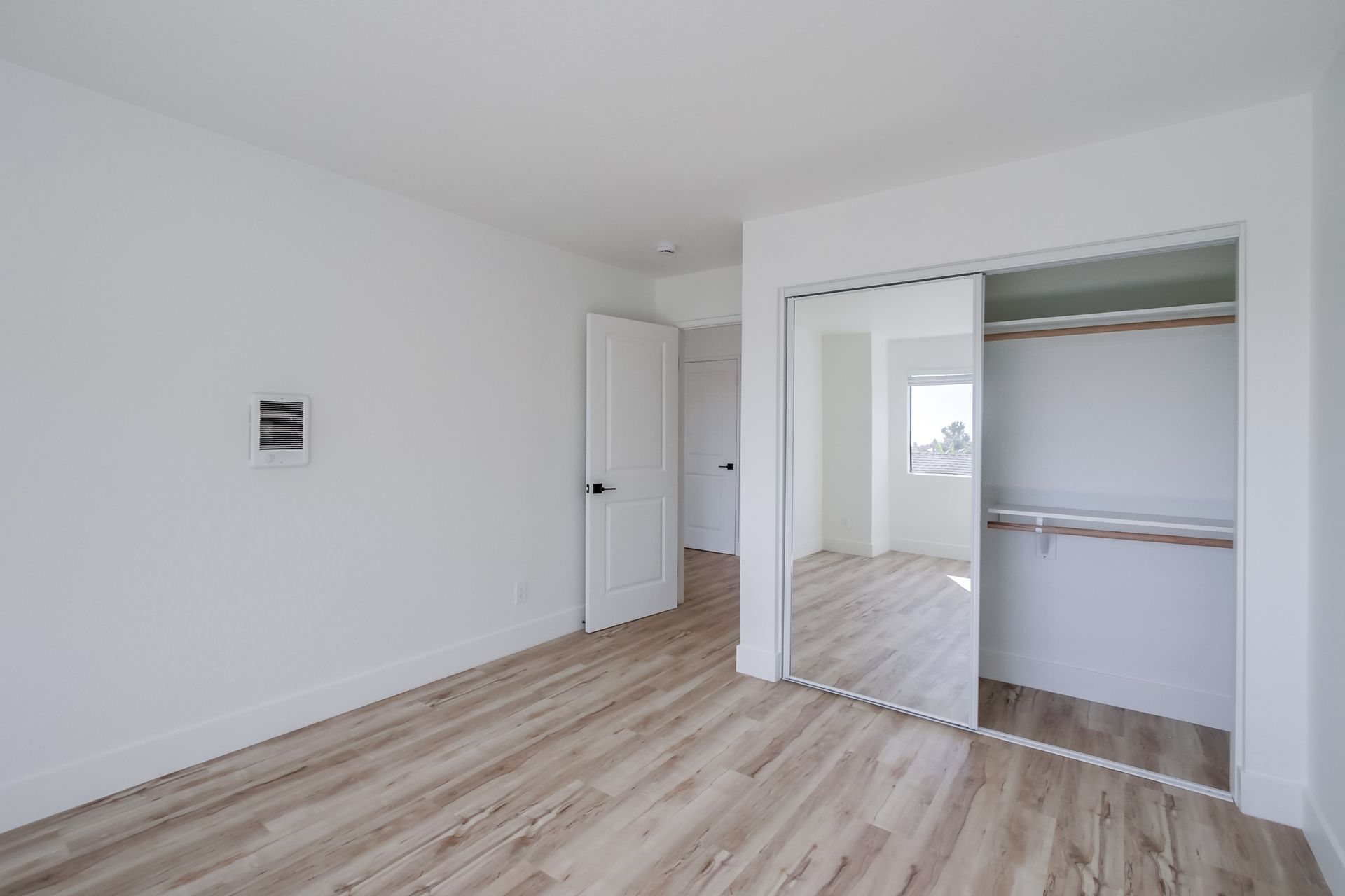 Empty bedroom with light wood floors, white walls, and mirrored closet doors.