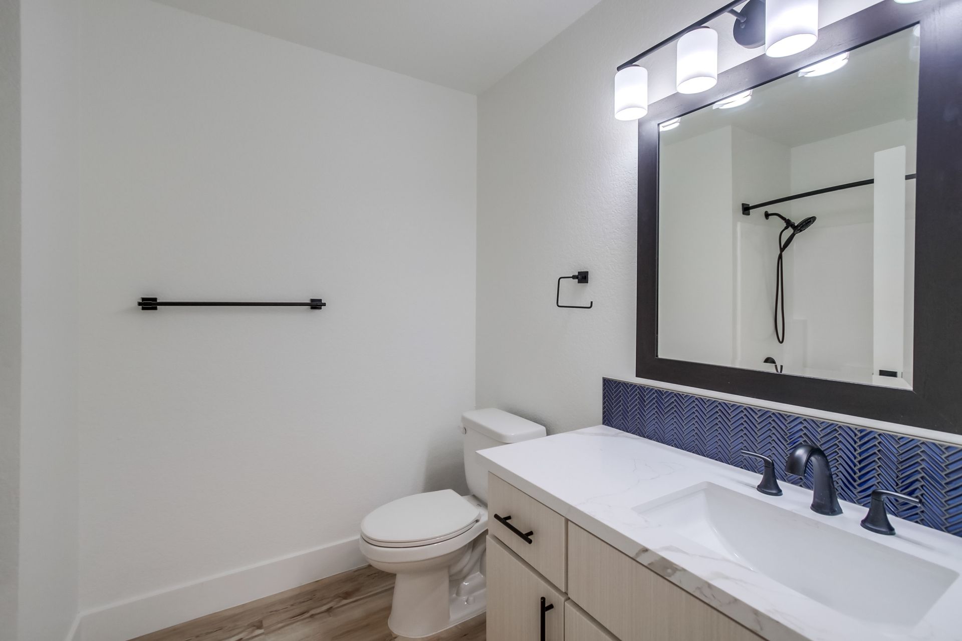 Bathroom with white walls, toilet, sink, and dark-framed mirror, blue mosaic tile backsplash, black fixtures.