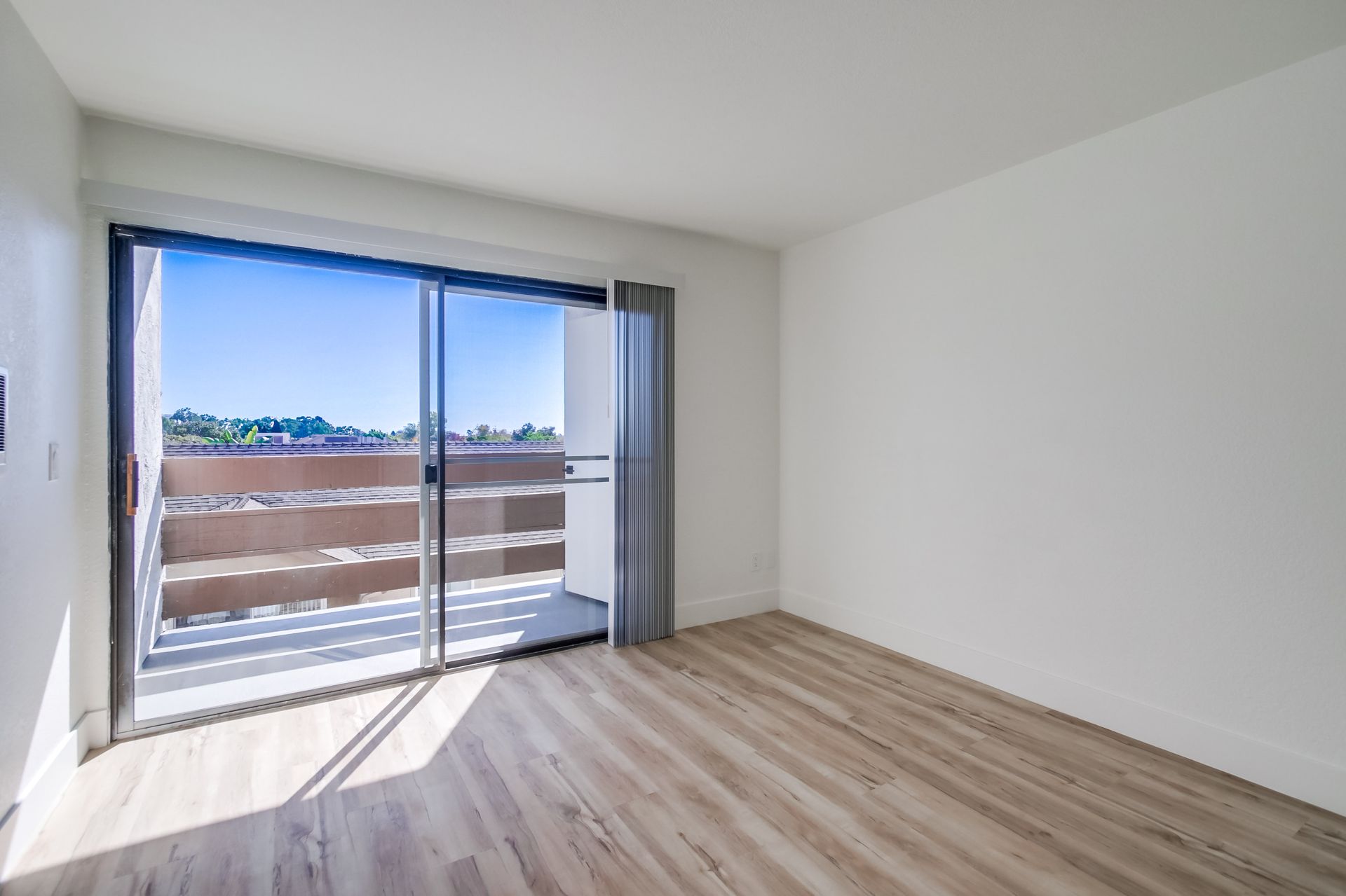 Empty room with sliding glass door to balcony. Light wood floors, white walls, and blue sky.