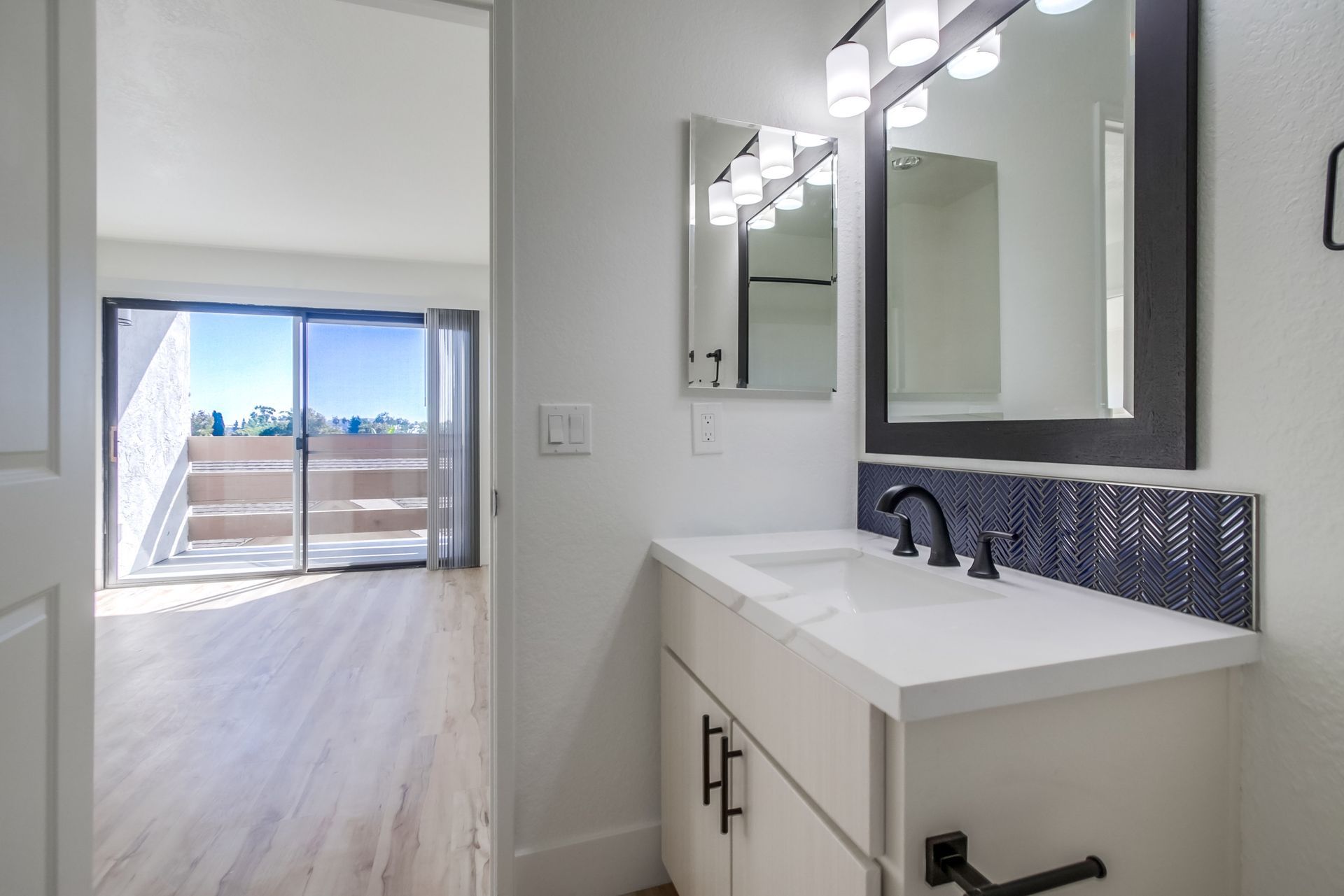Bathroom with white vanity, black-framed mirror, and blue tiled backsplash. View of a balcony through an open door.