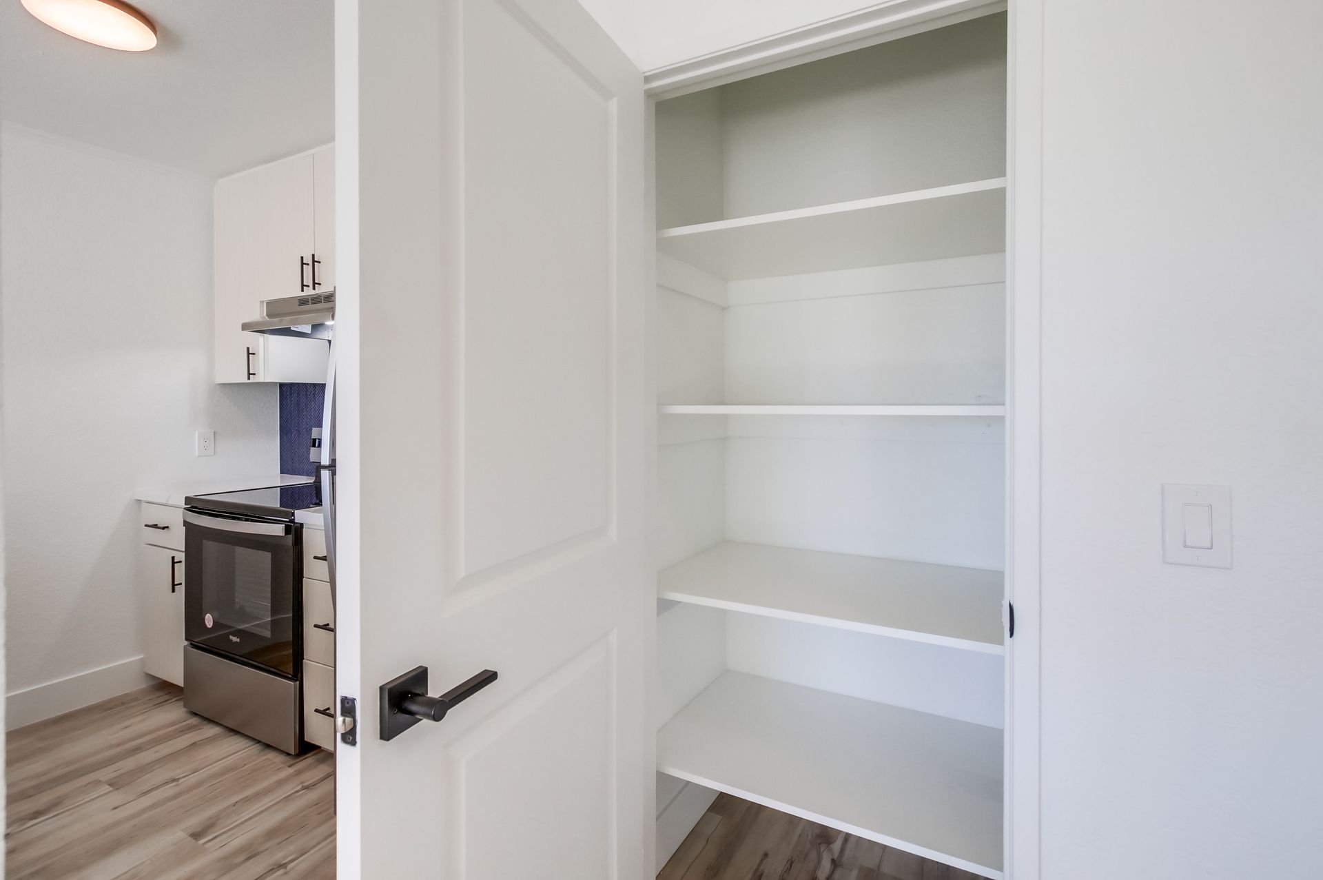 White pantry with open door, revealing empty shelves, next to a small kitchen.