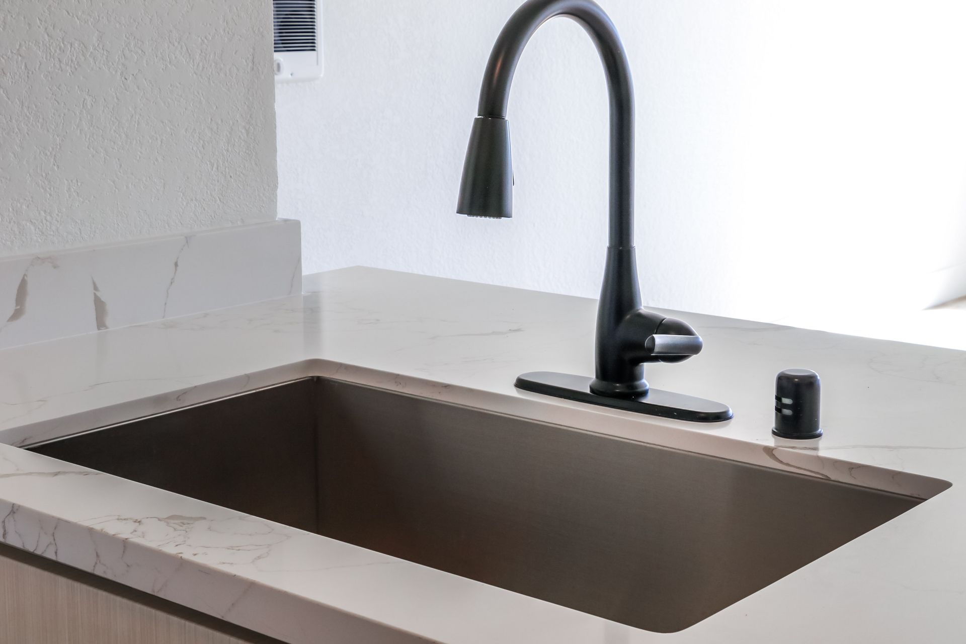 Black faucet over a square, dark-colored sink in a kitchen with a light-colored countertop.