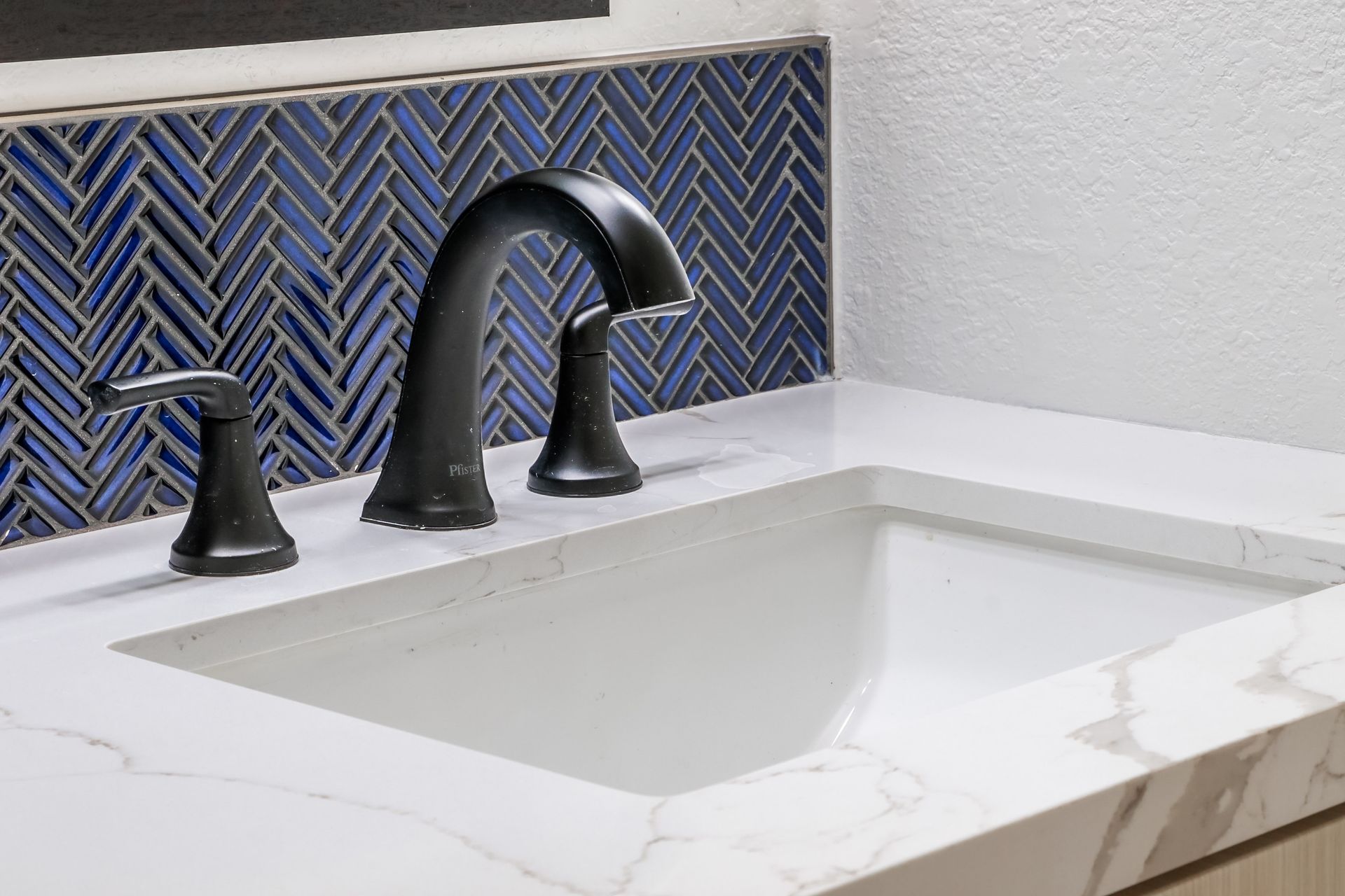 Close-up of a bathroom sink with dark fixtures, blue tile backsplash, and white countertop.