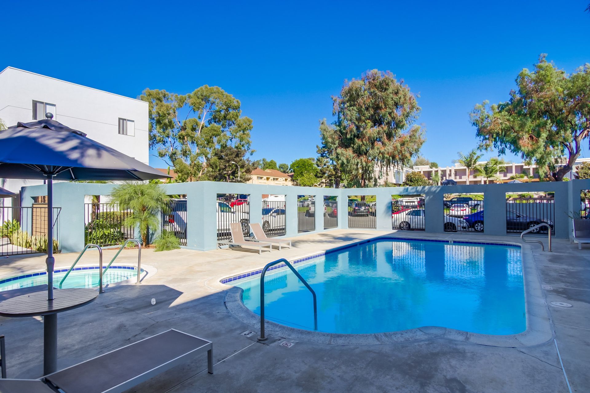 A pool surrounded by concrete and a light blue wall. A black umbrella and lounge chairs are by the pool.
