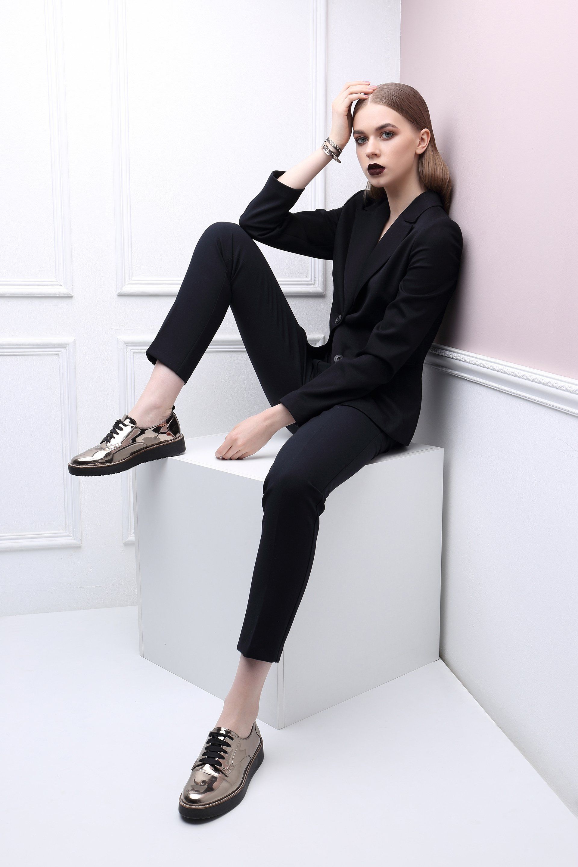 A woman in a black suit is sitting on a white cube.