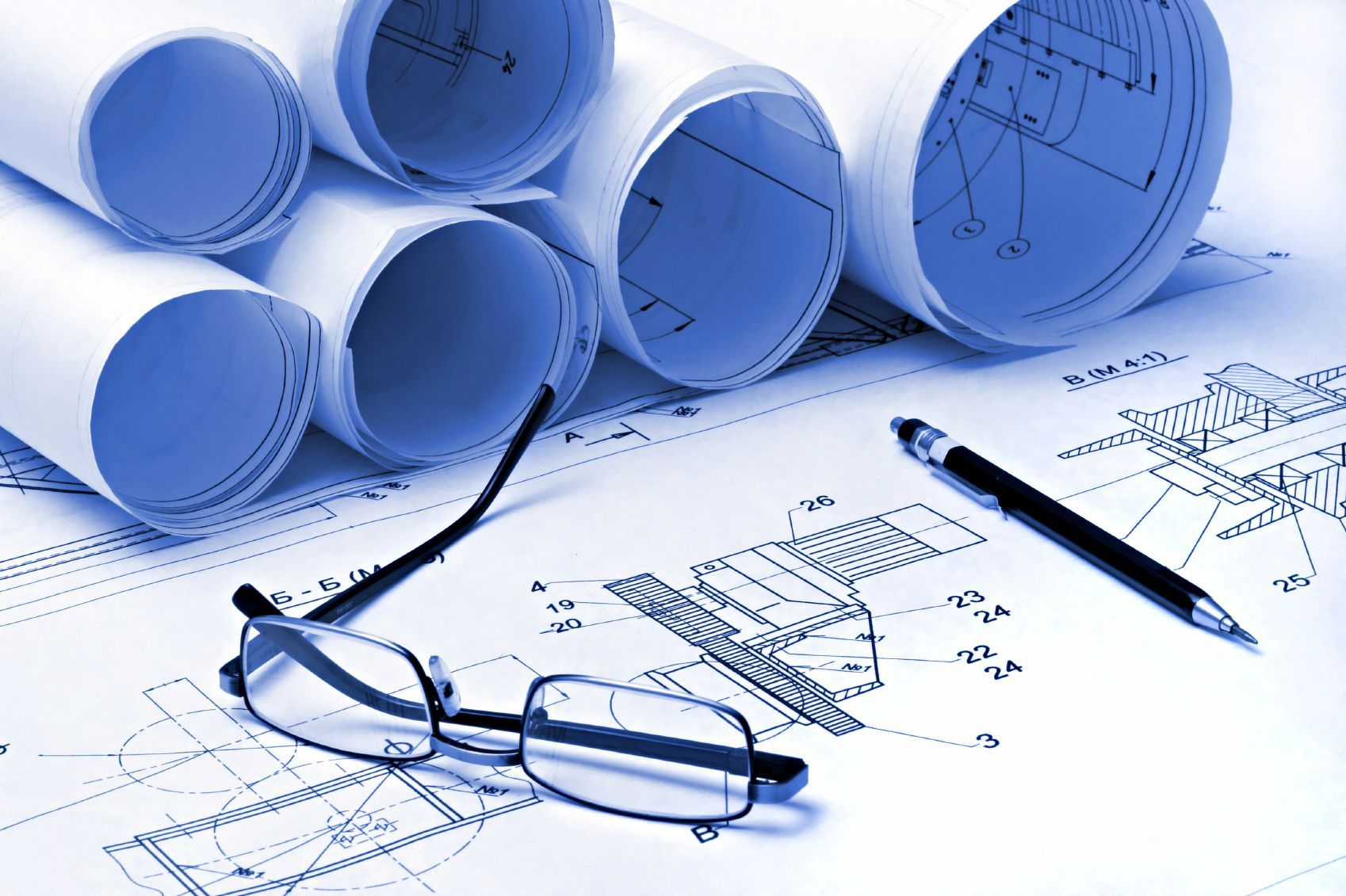 Blue-toned architectural blueprints and rolled plans; glasses and pen rest on the open documents.
