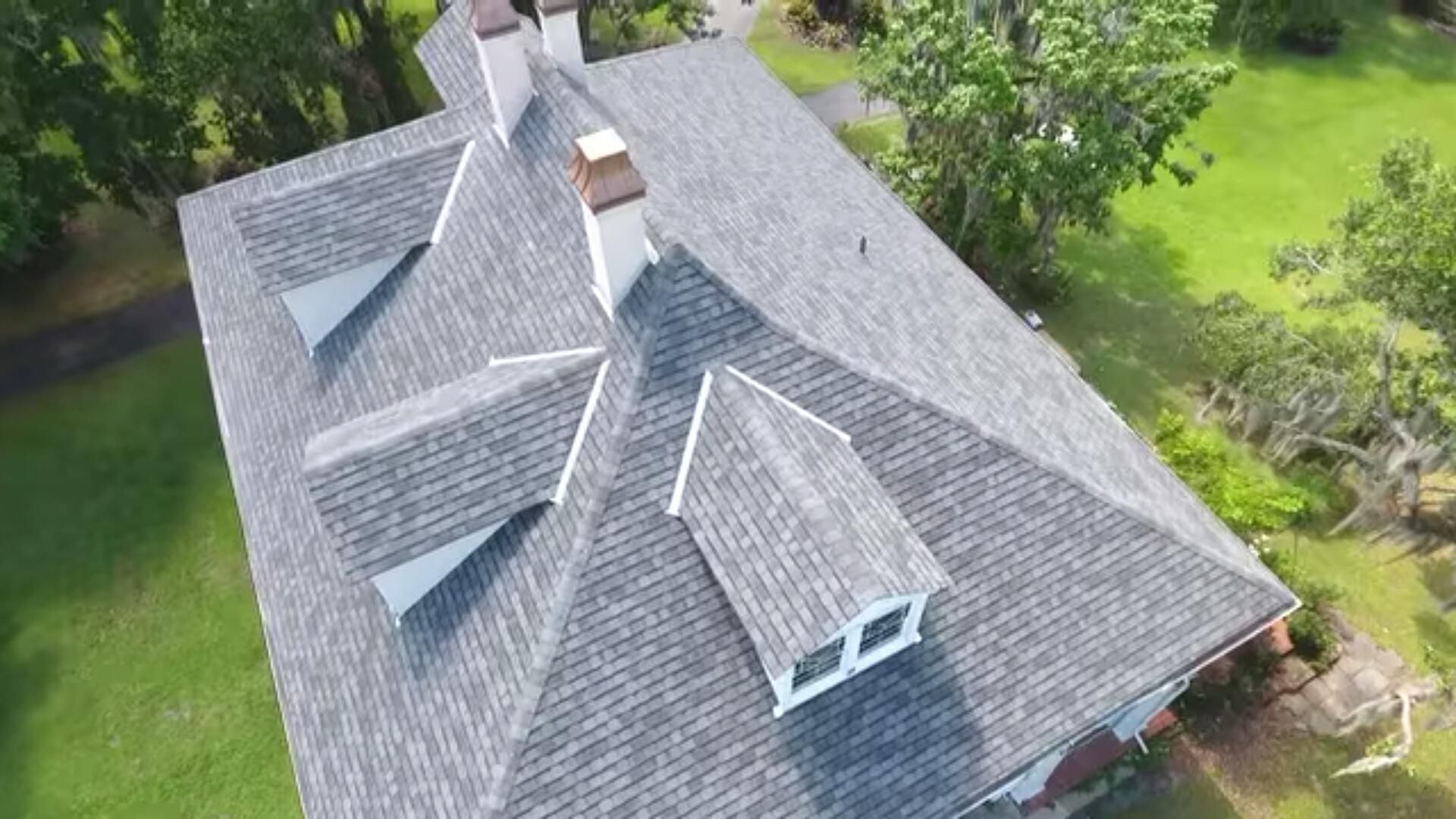 Aerial view of a house with a gray shingled roof, dormers, and chimneys, surrounded by green grass and trees.