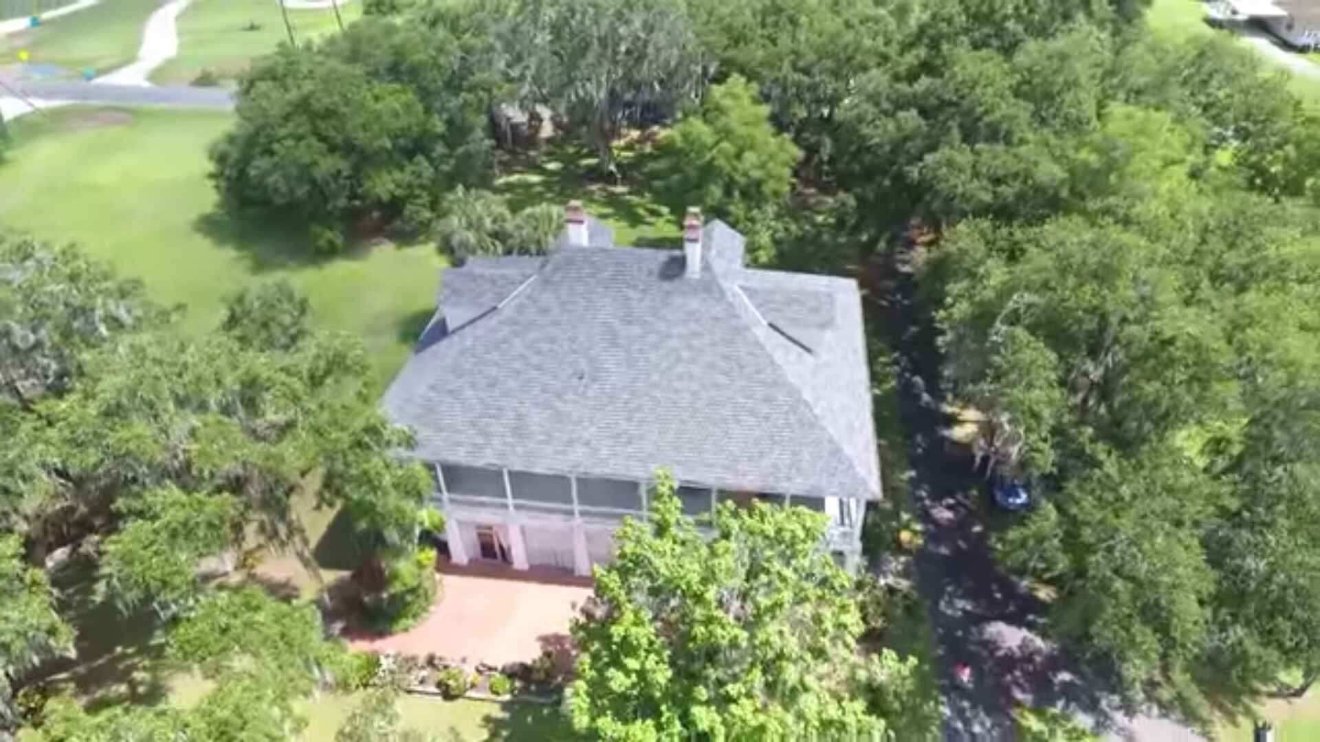 Aerial view of a large, white house with a gray roof surrounded by green trees and a red brick porch.