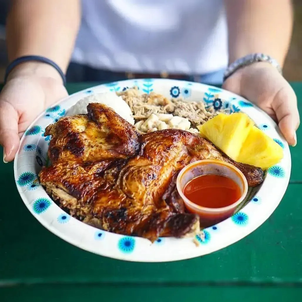 Person holds plate of grilled chicken, rice, macaroni salad, pineapple, and red sauce.