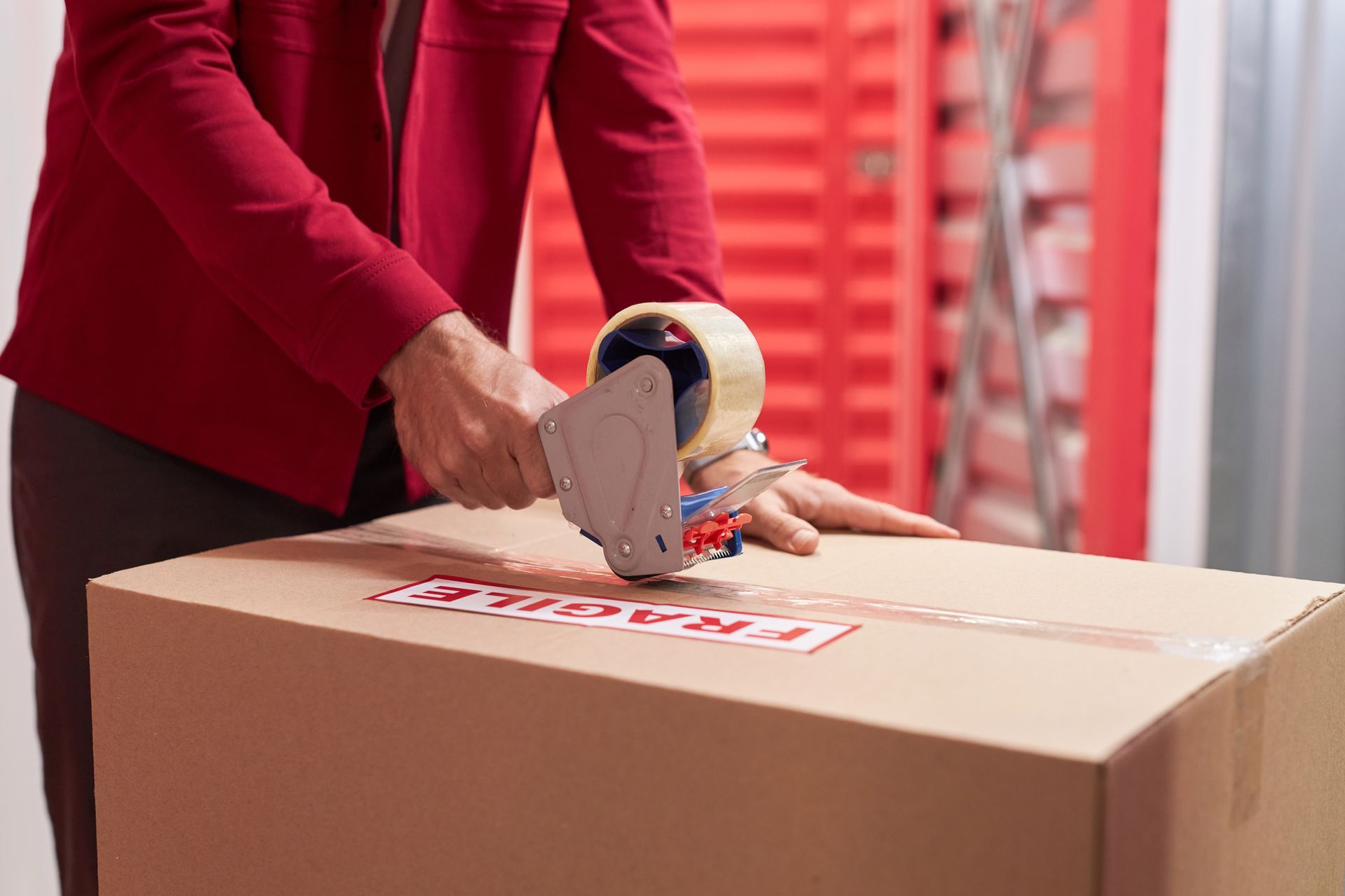 Person in a red blazer using a tape gun to seal a cardboard box marked fragile near storage units. Person in a red blazer using a tape gun to seal a cardboard box marked fragile near storage units.