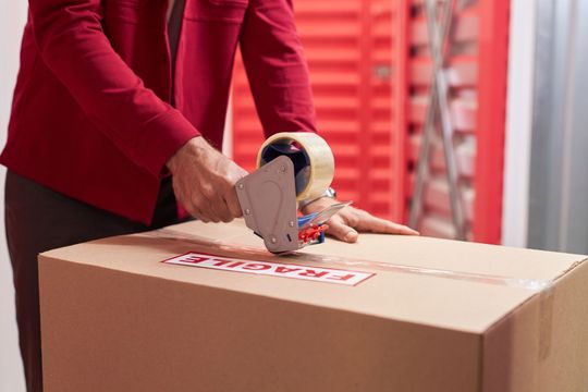 Person in a red blazer using a tape gun to seal a cardboard box marked fragile near storage units. Person in a red blazer using a tape gun to seal a cardboard box marked fragile near storage units.