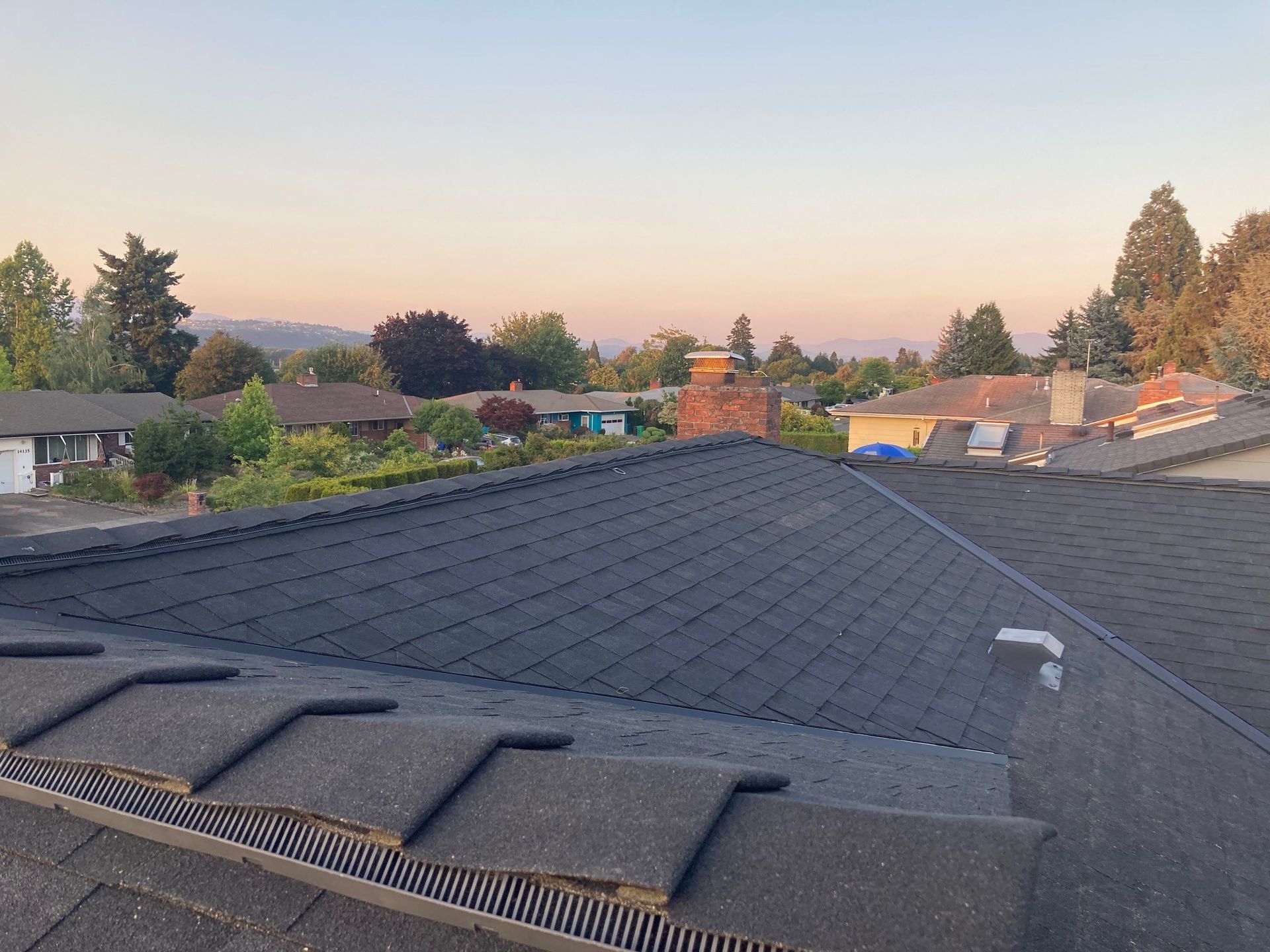 Rooftop view of a residential area at sunset, showing asphalt shingles and a brick chimney. Trees and mountains are in the distance.