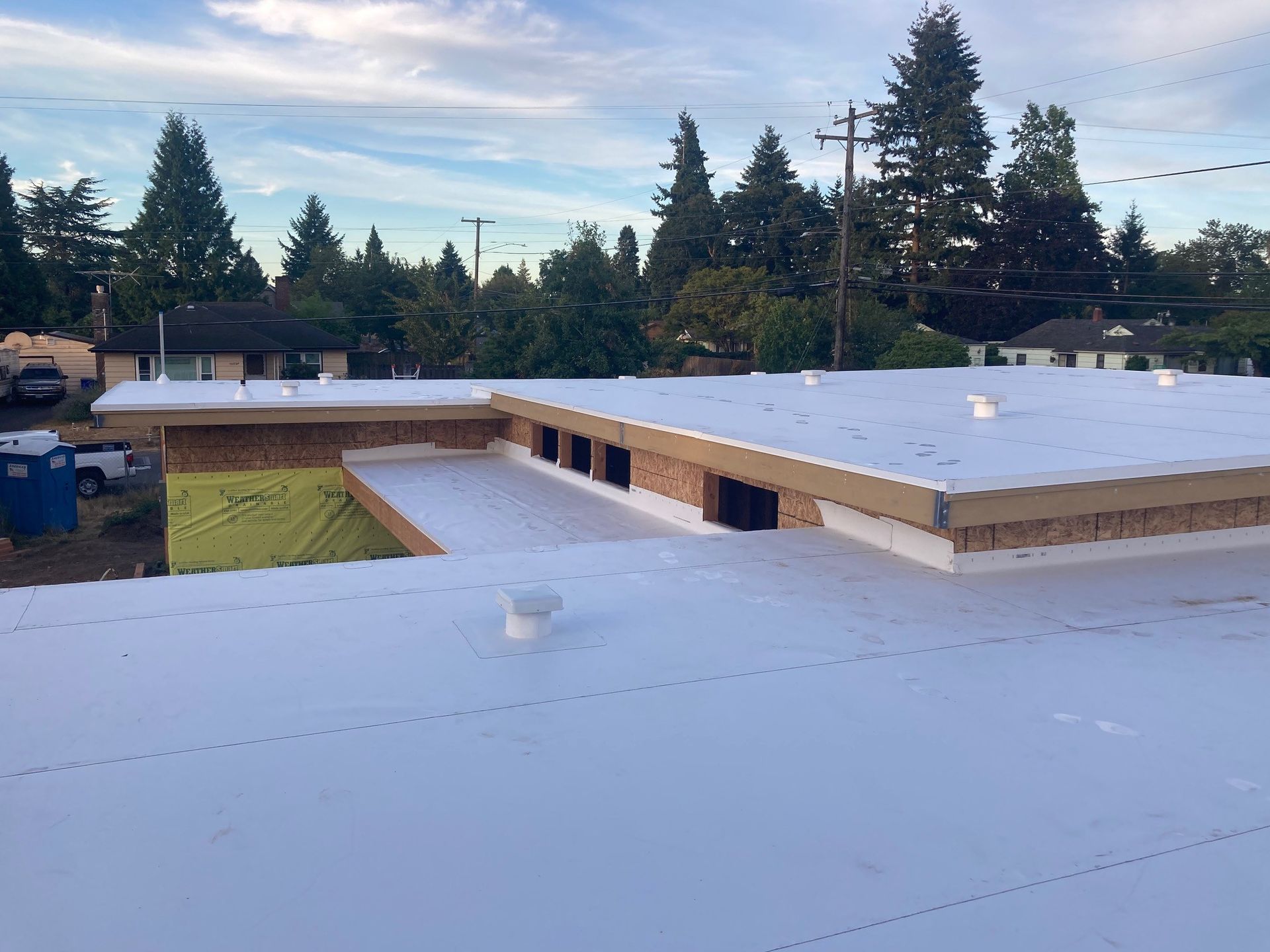 Flat roof under construction with white membrane, vents, and exposed wood framing.
