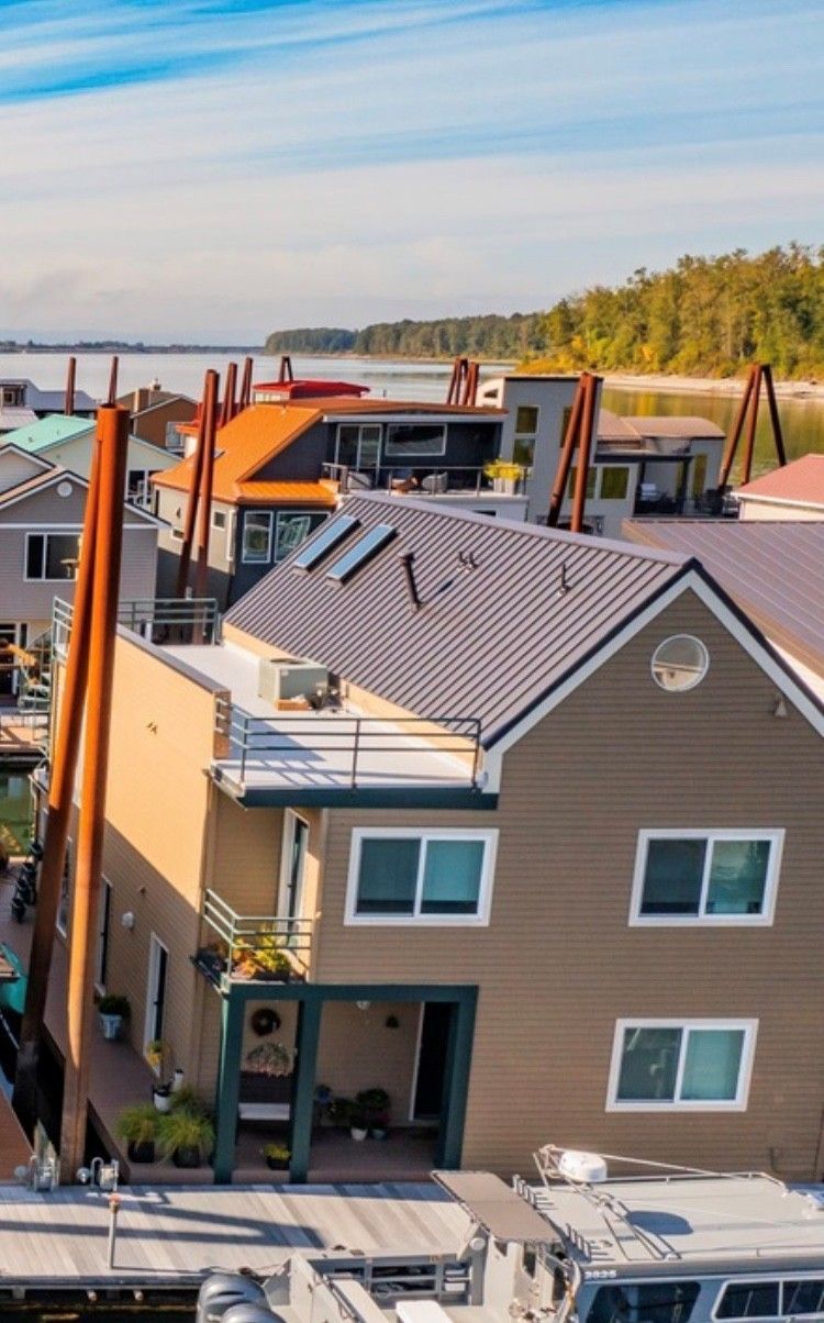 Floating homes on a lake, with brown, orange, and white exteriors. Wooden pilings support the structures; blue sky.