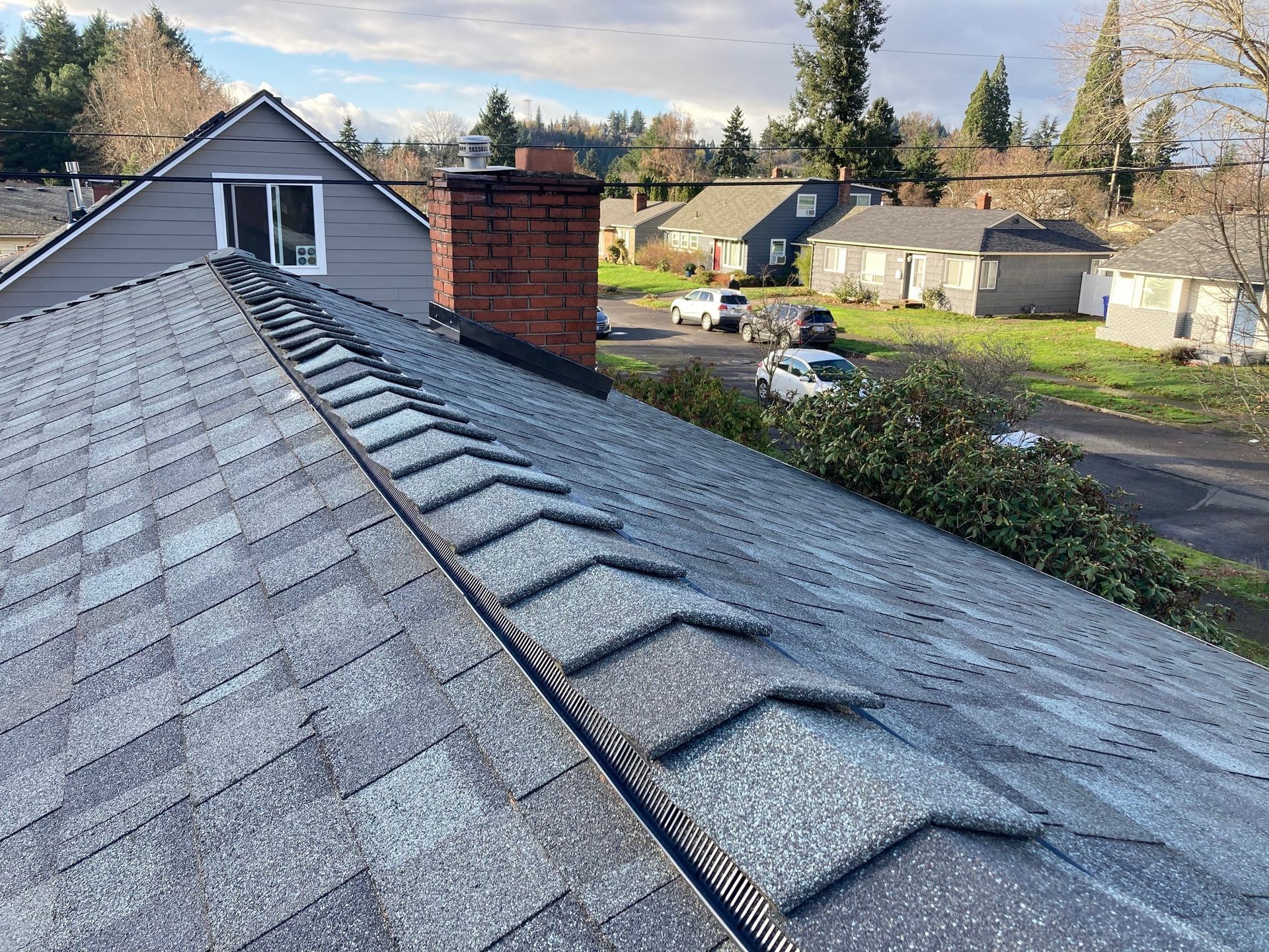 Rooftop with gray shingles, a brick chimney, and a view of houses and trees on a sunny day.