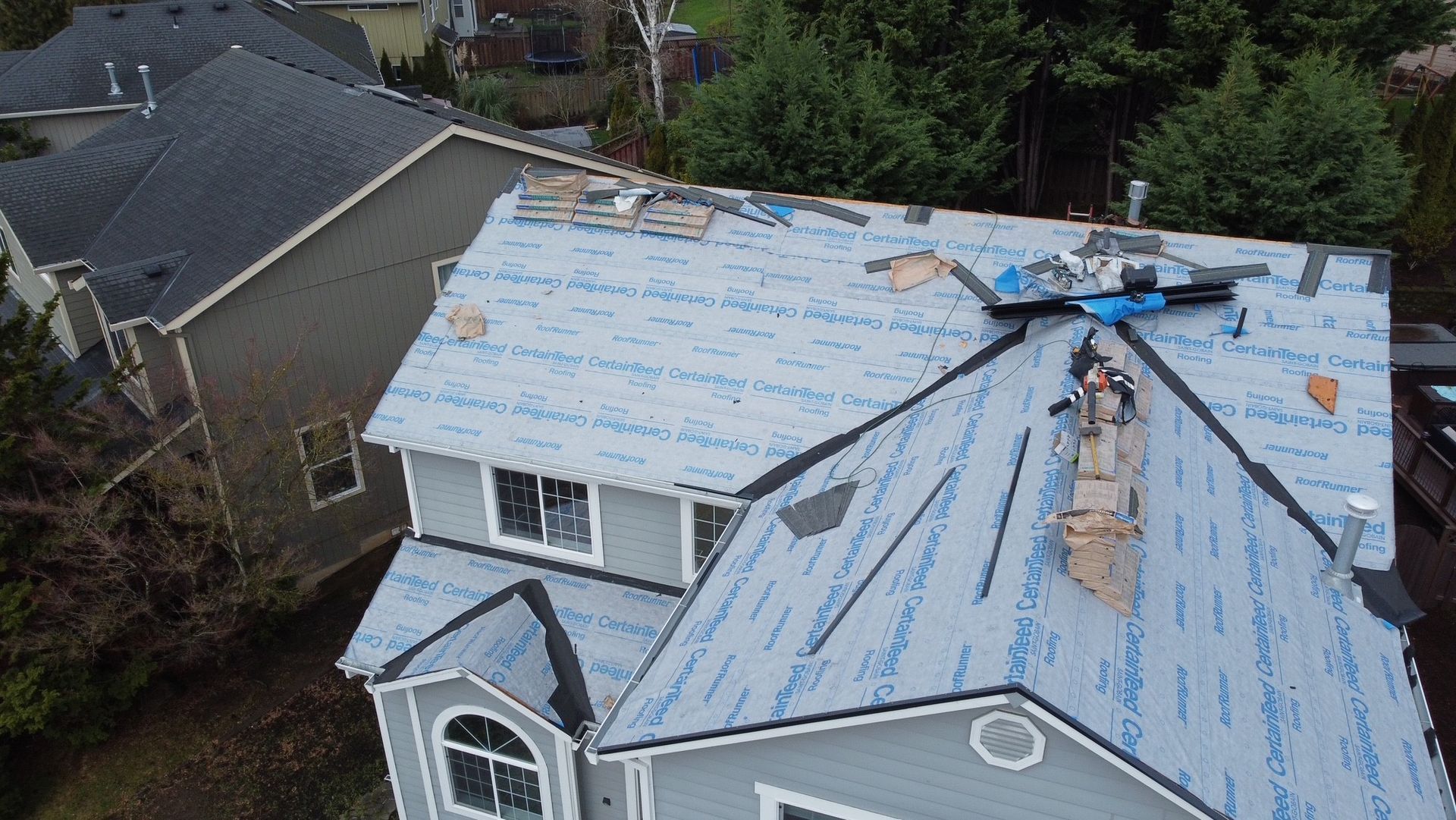 Aerial view of a house with a partially completed roof replacement, blue underlayment visible, surrounded by trees.