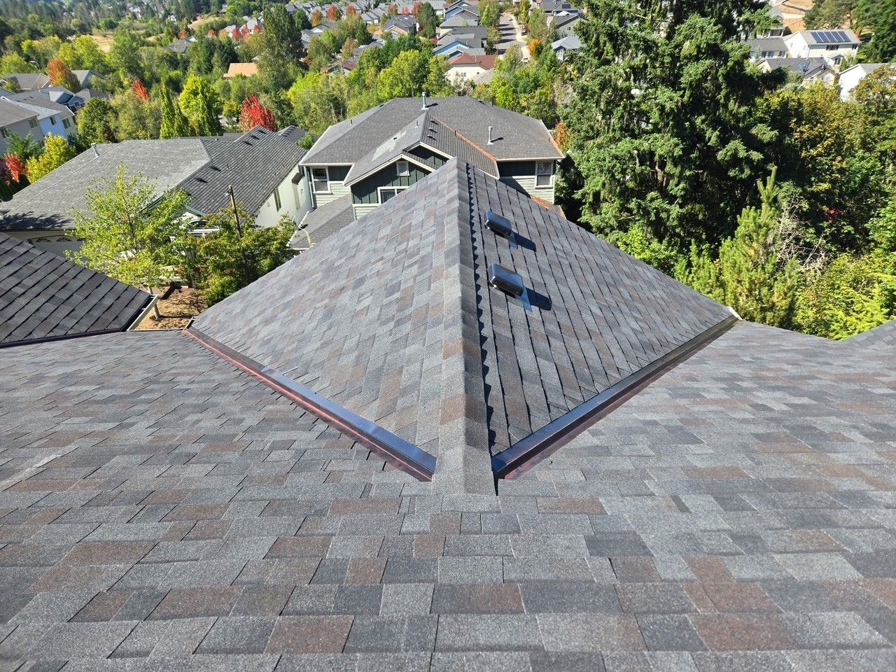 Close-up of a house roof with dark gray shingles, with other homes and green trees in the background. Close-up of a house roof with dark gray shingles, with other homes and green trees in the background.