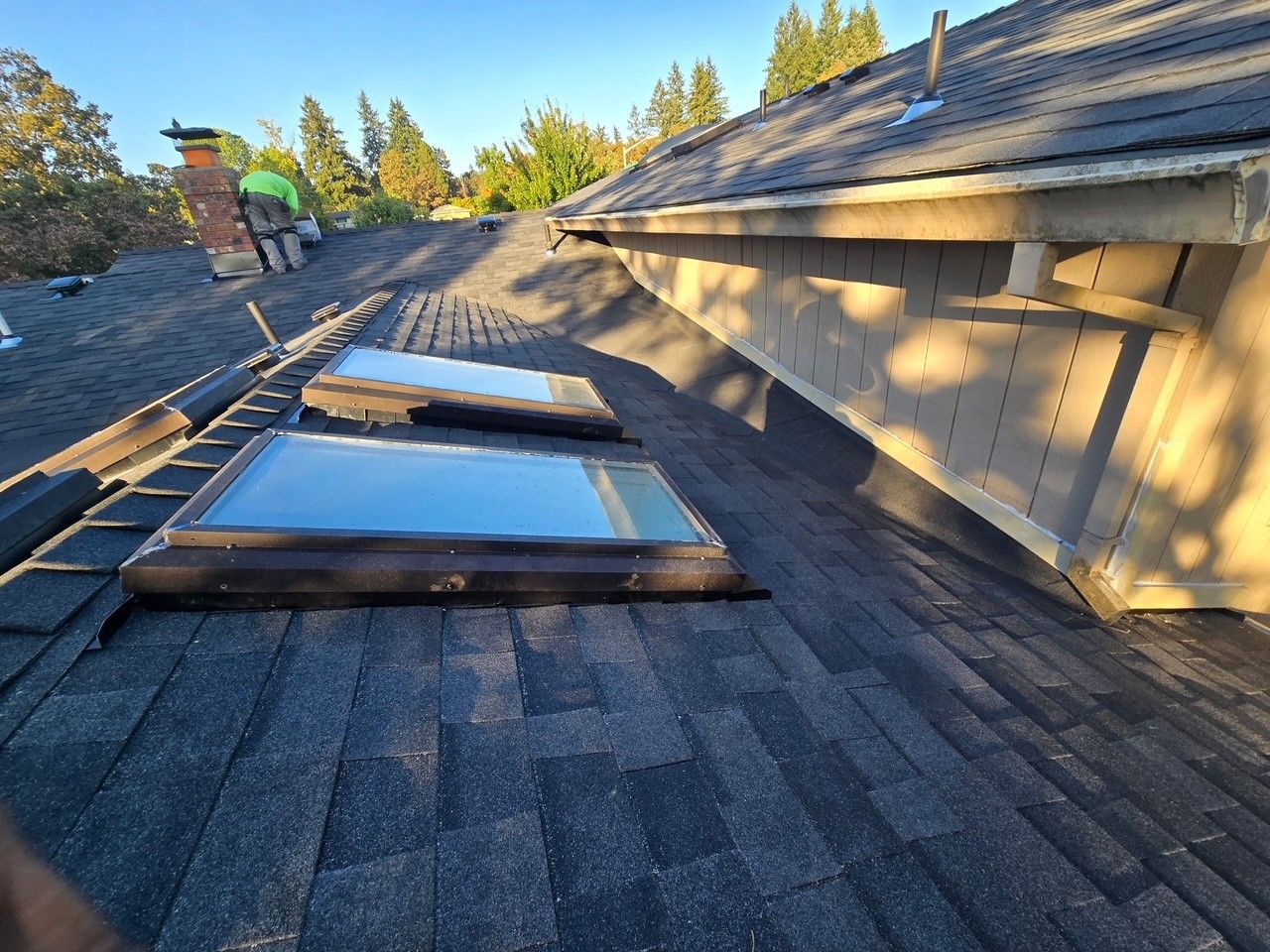 A roof with two skylights. Dark shingles, building with beige siding, and a person working in the background.