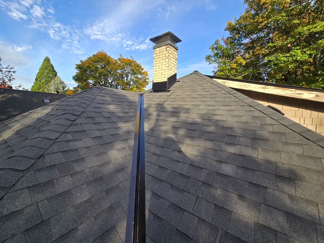 View of a dark gray shingled roof with a chimney and trees against a blue sky.