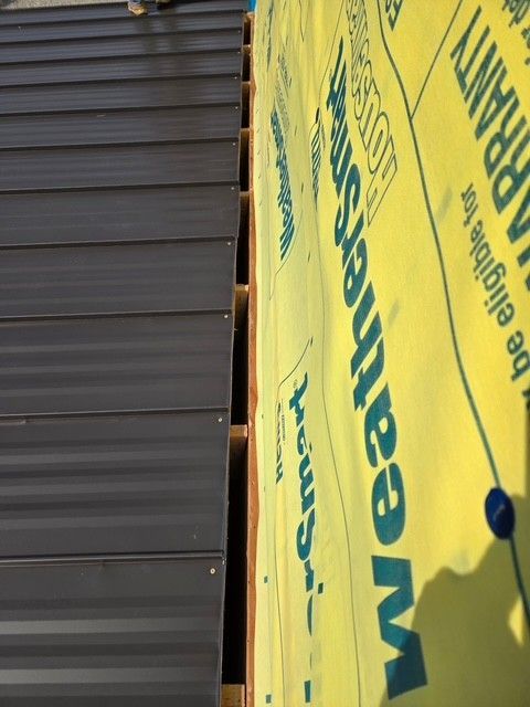 Close-up of a dark metal roof edge next to yellow WeatherSmart house wrap and exposed wooden beams.