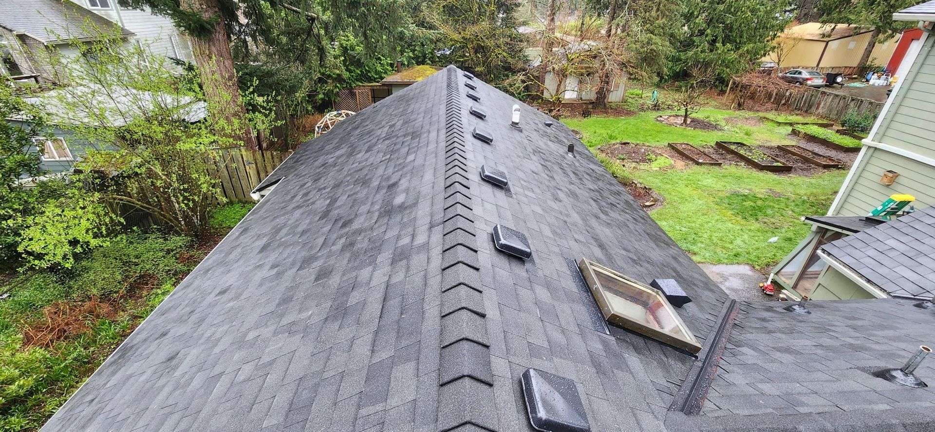 Overhead view of a gray asphalt shingle roof with a ridge vent, trees, and a garden.