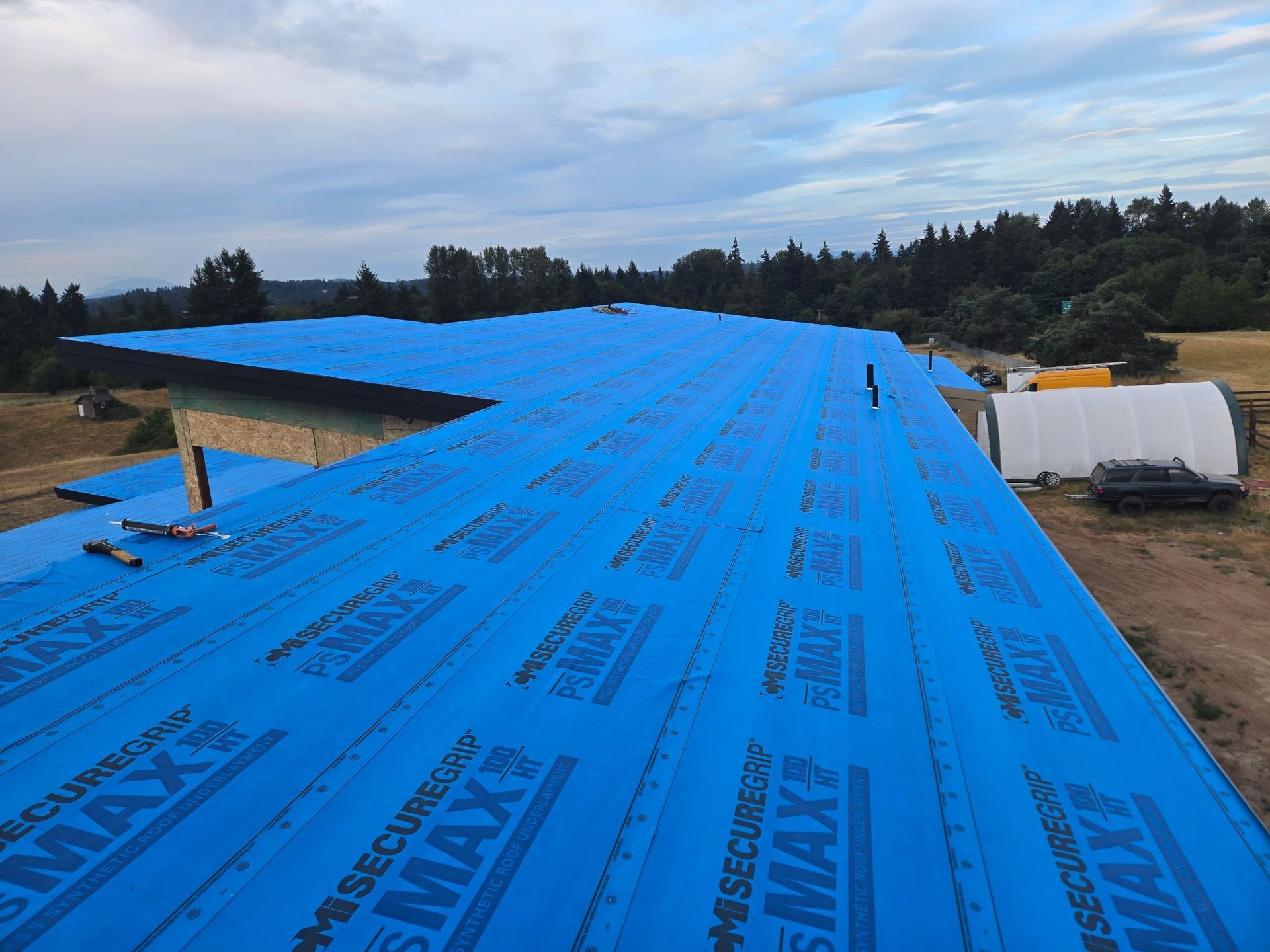 Blue roofing material covers a large roof of a building under construction, rural setting.