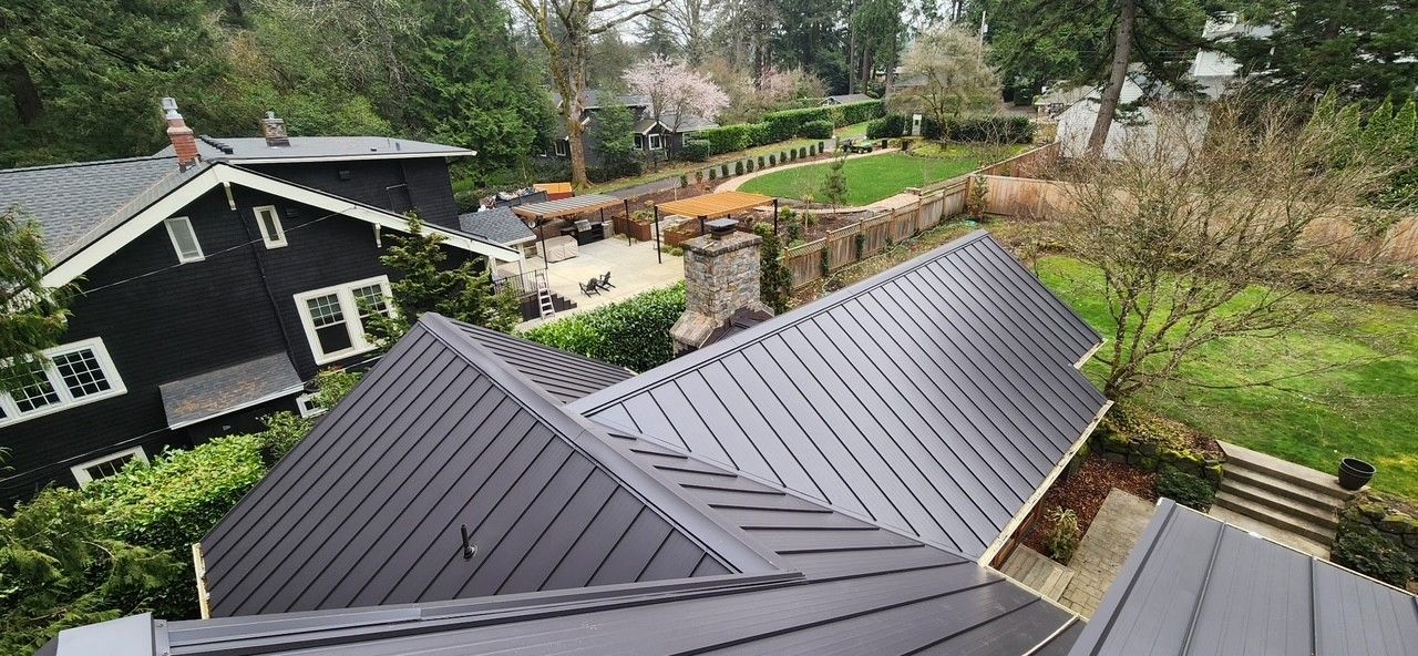 Overhead view of a house with a dark metal roof. A second house and a grassy yard with stone walls are in the background.