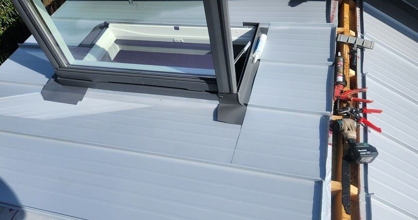 Skylight installation on a gray metal roof, with tools and open window.