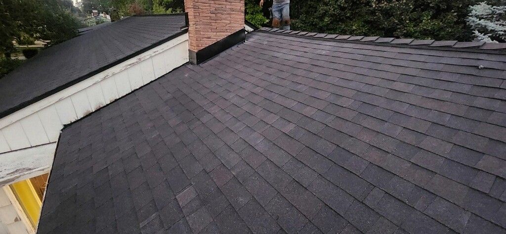 A dark asphalt shingle roof. A brick chimney stands atop the roof with a person standing by it.