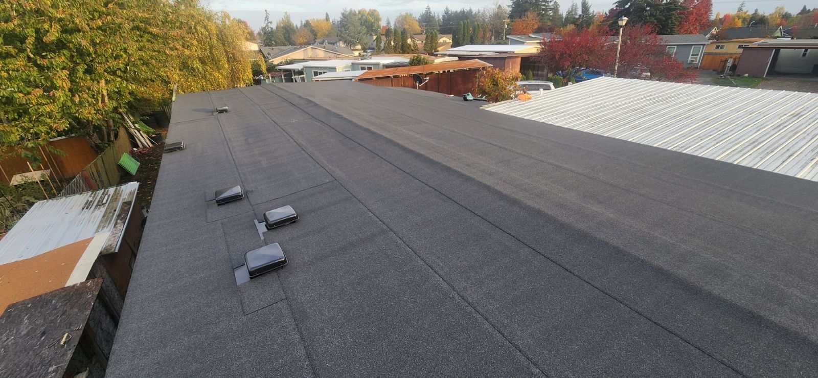 View of a flat, dark gray roof with several vents, trees and buildings in the background. Overcast sky.