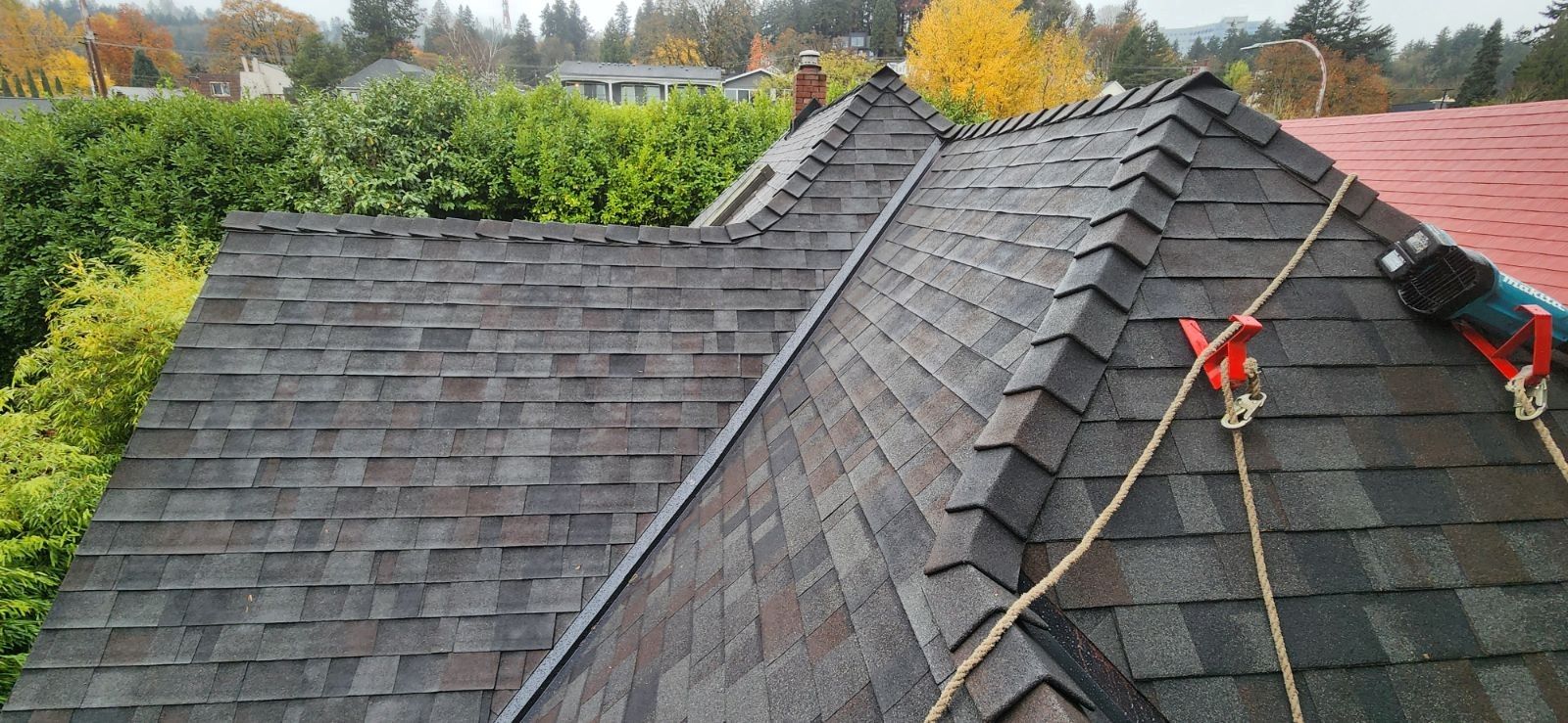 Roofer on a residential roof, wearing safety gear. Shingles are gray, setting is overcast. Safety ropes present.