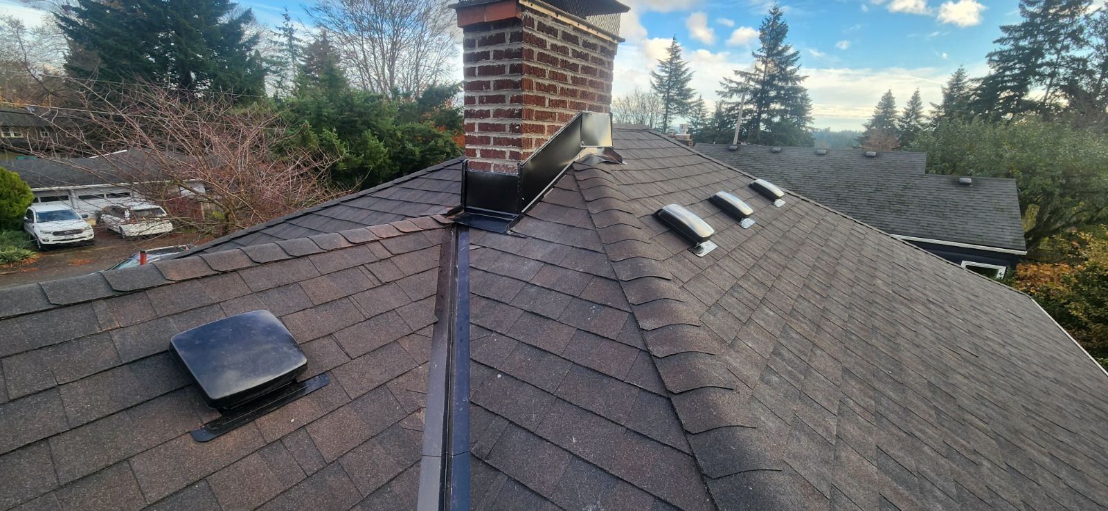 A close-up view of a roof with shingles, a brick chimney, and vents against a partly cloudy sky.