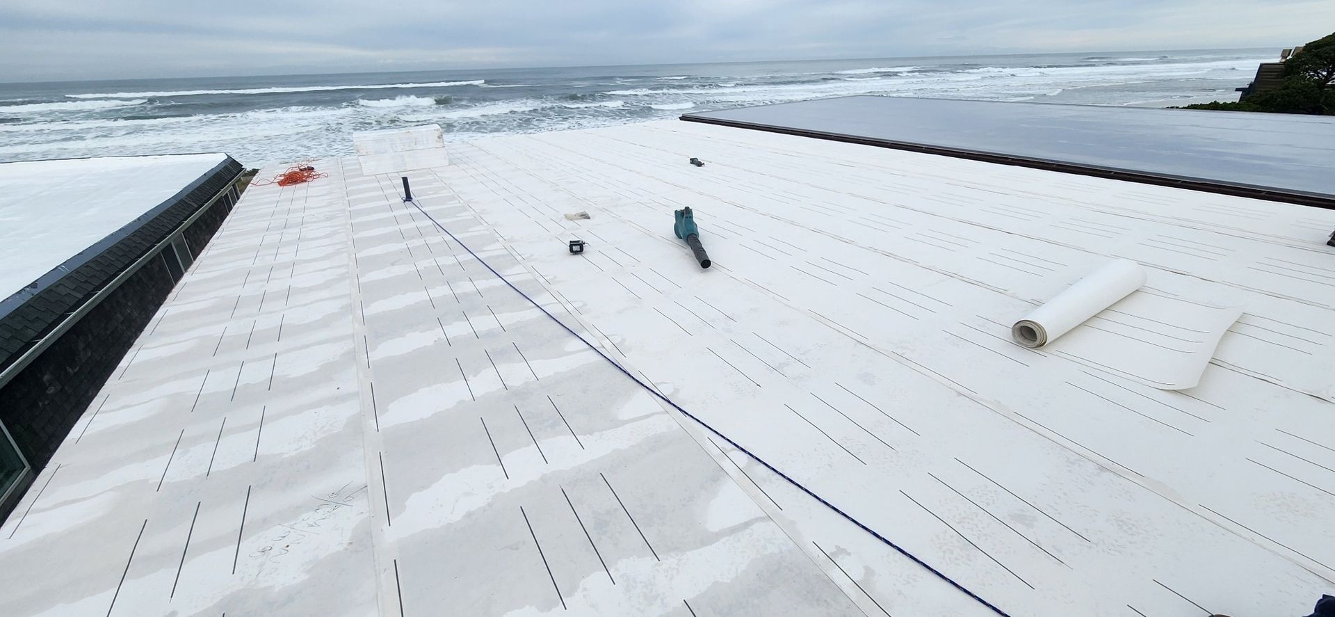 Workers on a white roof by the ocean. One worker in green suit. Waves crash in the background. Cloudy sky.
