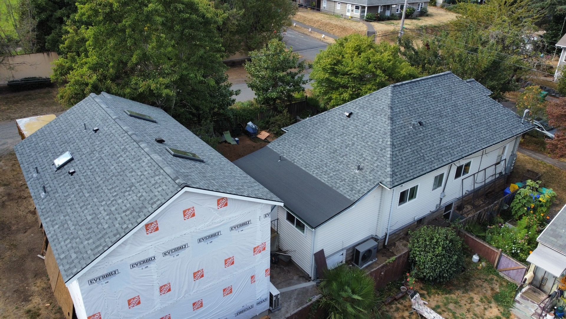 Aerial view of two houses with new gray roofs, one under construction, surrounded by trees.