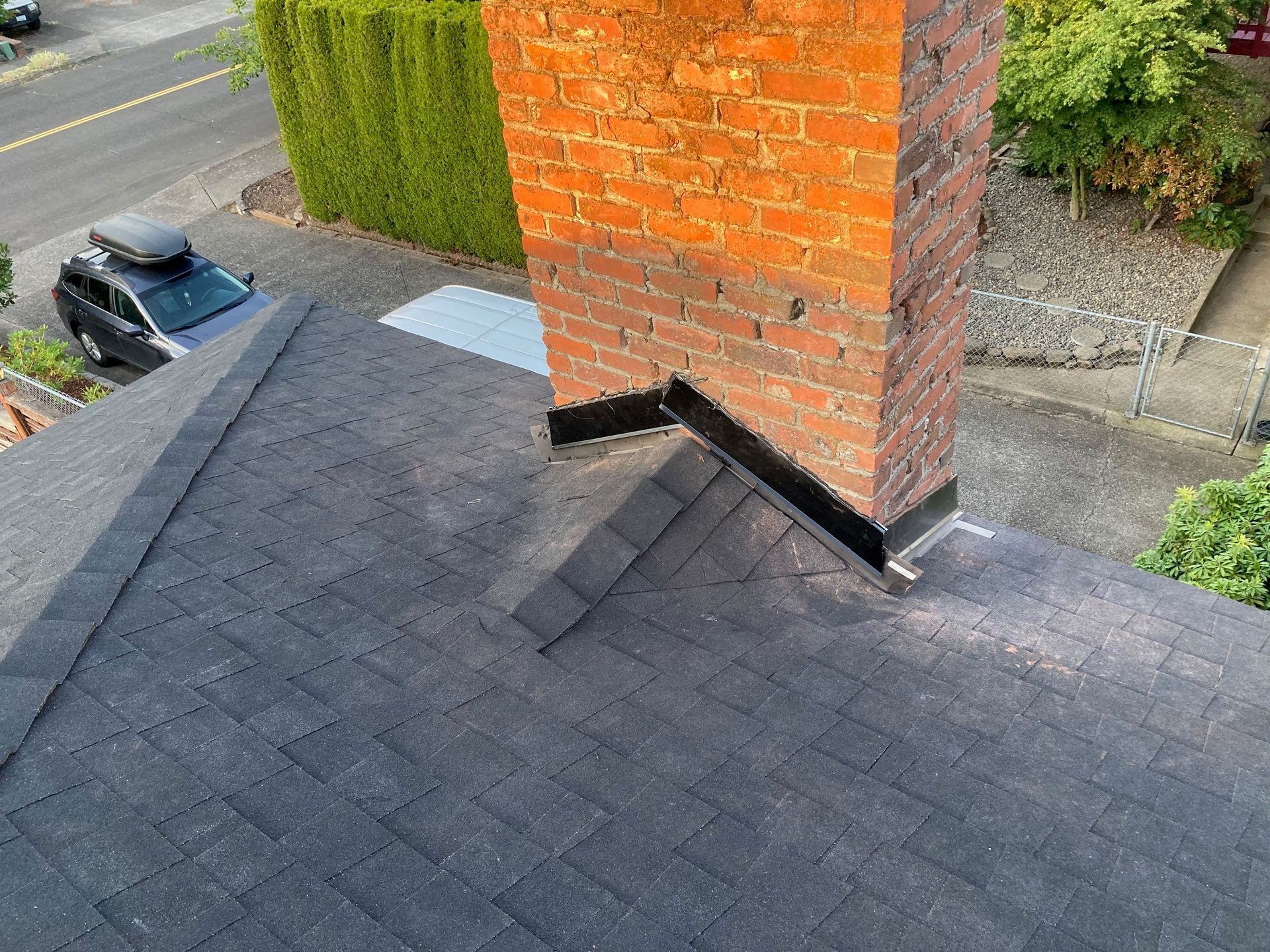 View of a brick chimney on a dark shingled roof, with flashing around the chimney base and a car parked in the street.