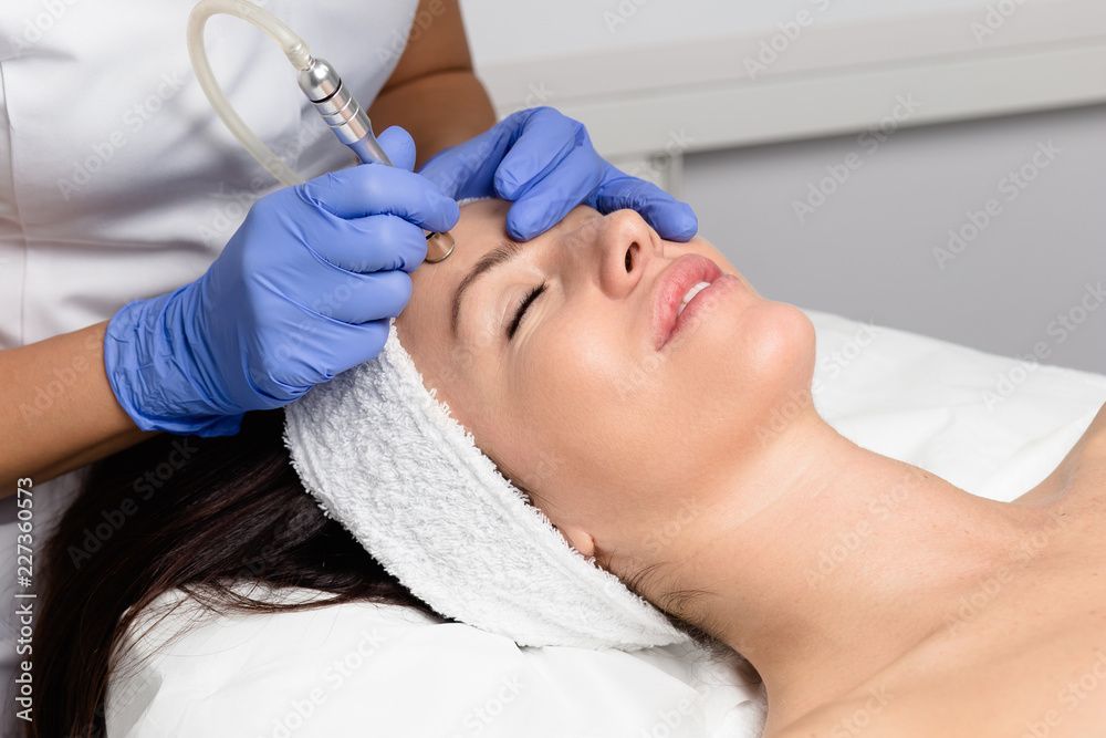 Woman receiving microdermabrasion treatment; esthetician using a wand on her forehead, wearing blue gloves.