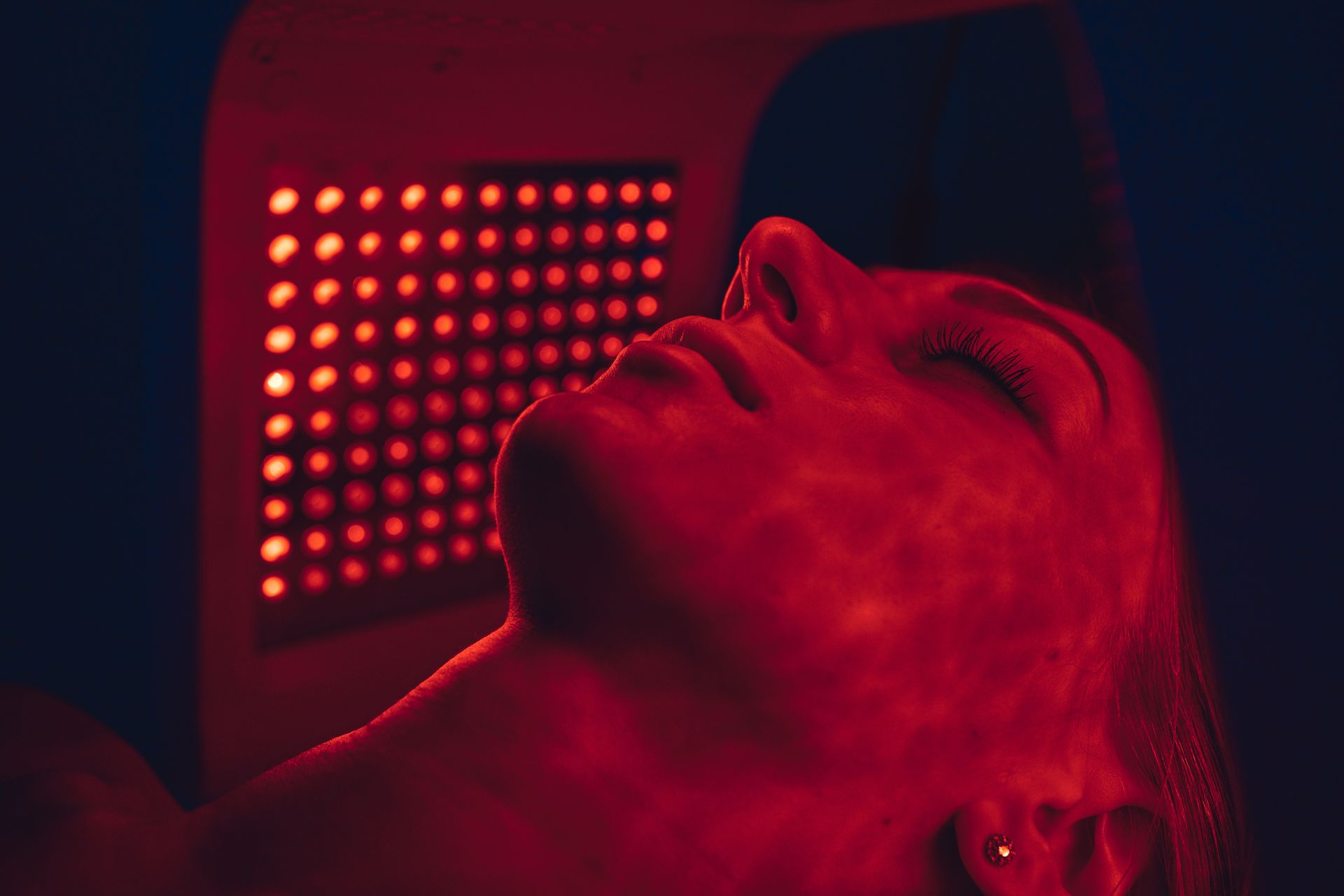 Woman's face illuminated by red light from a skincare LED panel. Eyes closed.