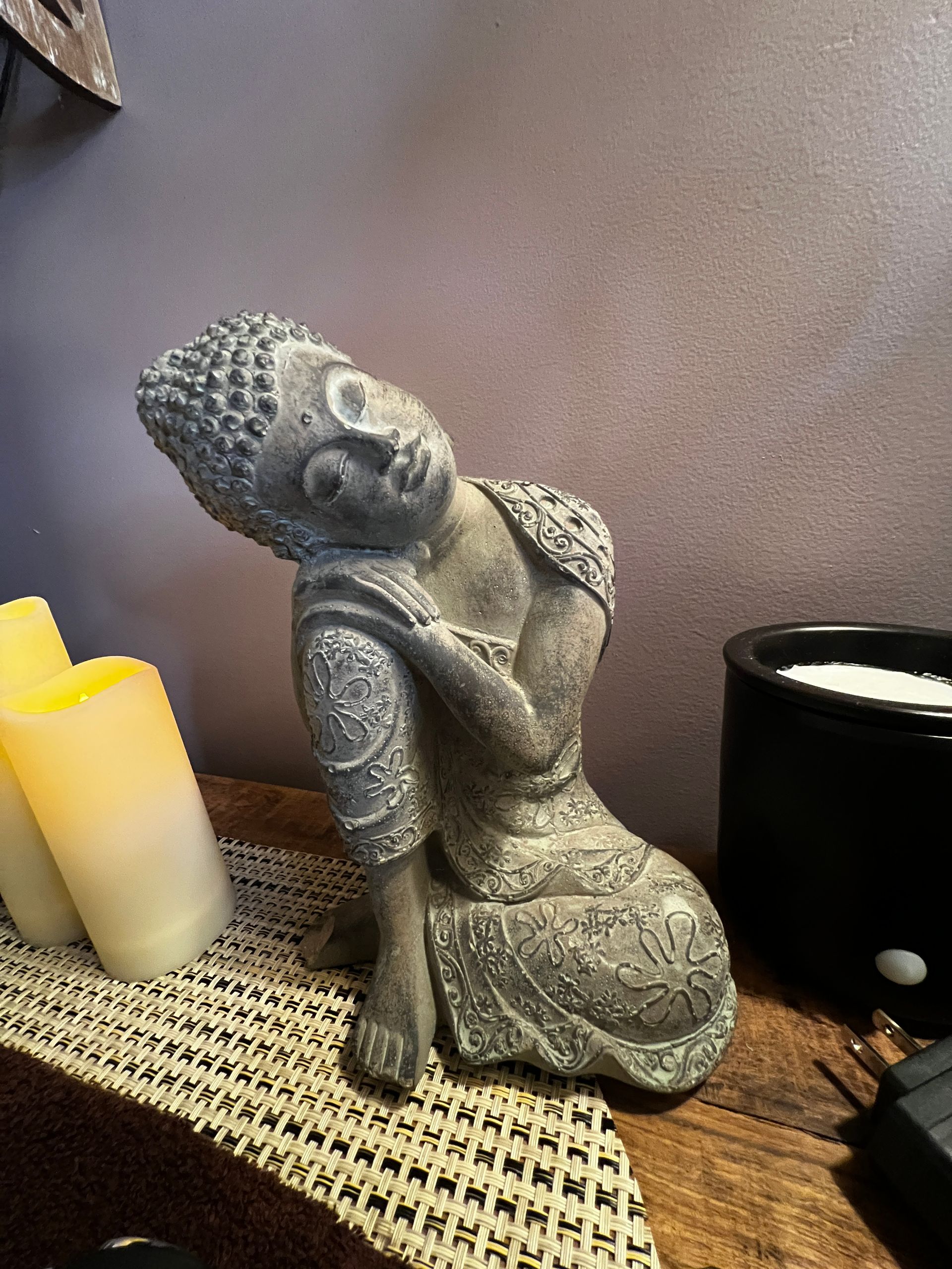 A seated grey Buddha statue with closed eyes, leaning against its knee, beside candles and a black pot.