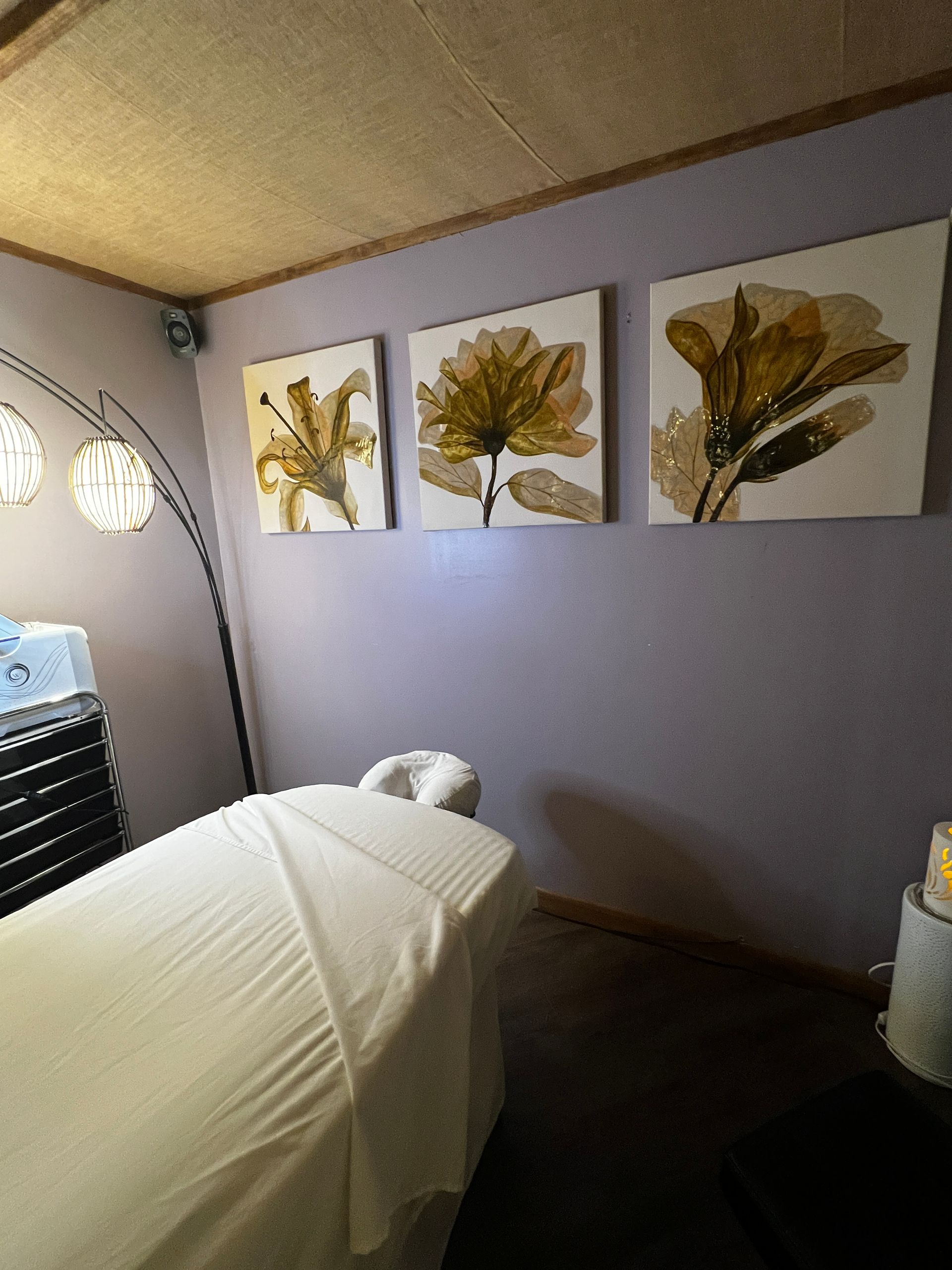Massage room: white-covered table, floral artwork, soft lighting, purple wall.
