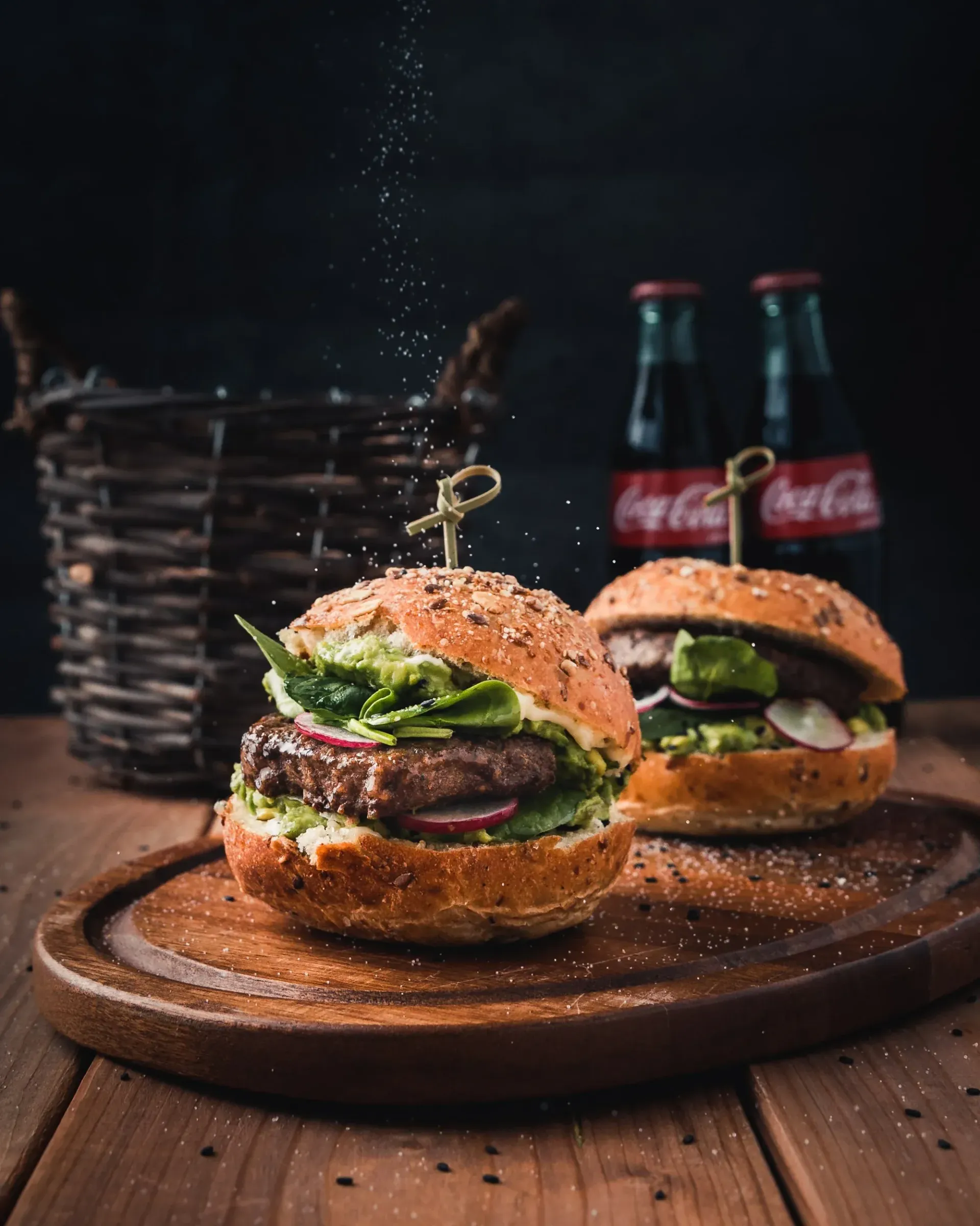 Two cheeseburgers on a wooden board with two Coca-Cola bottles in the background