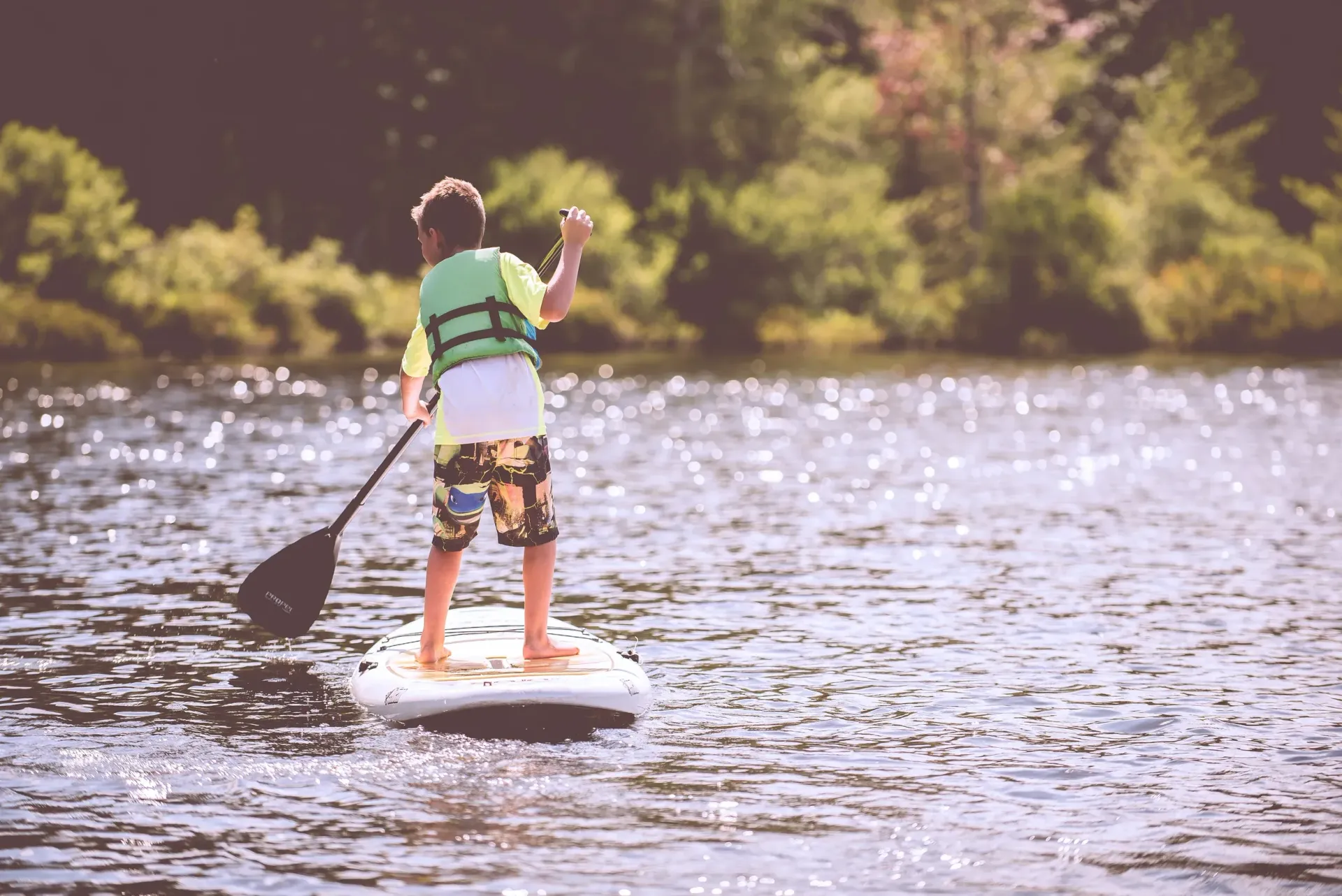 Person paddleboarding on a river, wearing a green shirt and patterned shorts, with trees in the background