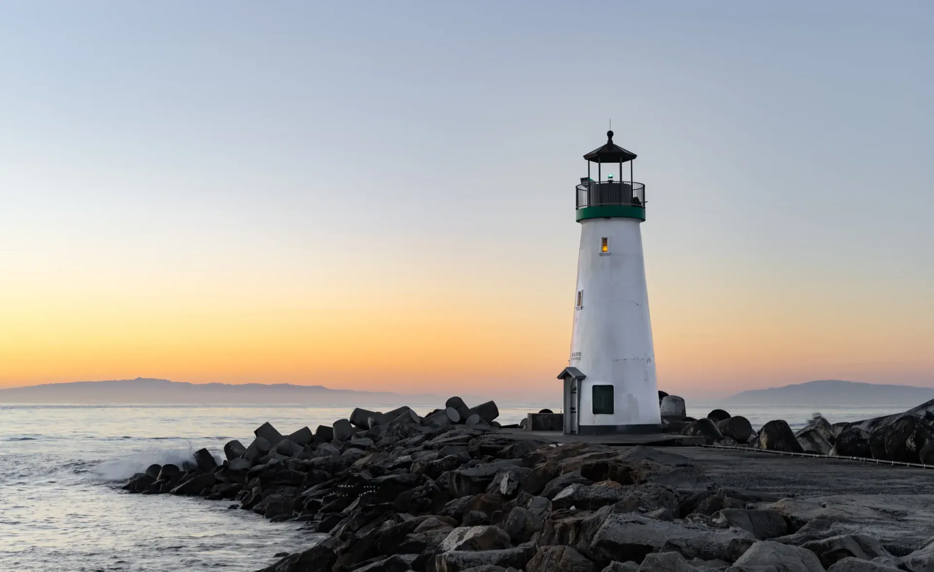 White lighthouse on rocky shore at sunset with calm water and pastel sky