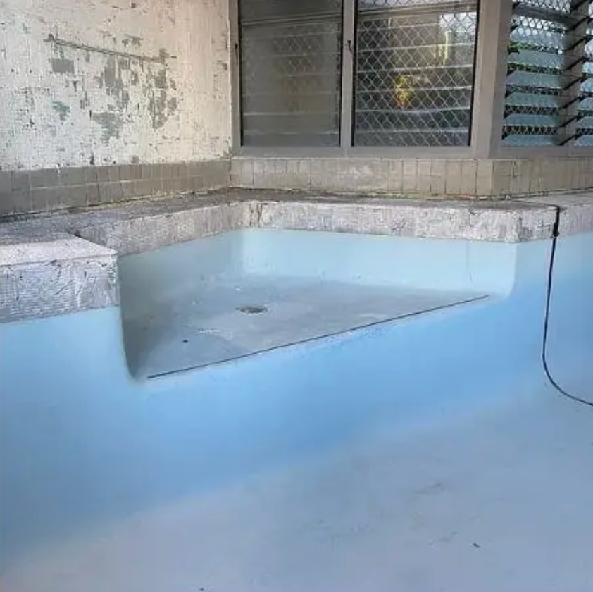 A light blue swimming pool corner bench submerged in water, set against a weathered wall and a window with a metal grate.  — East Coast Pool Resurfacing In Port Macquarie, NSW